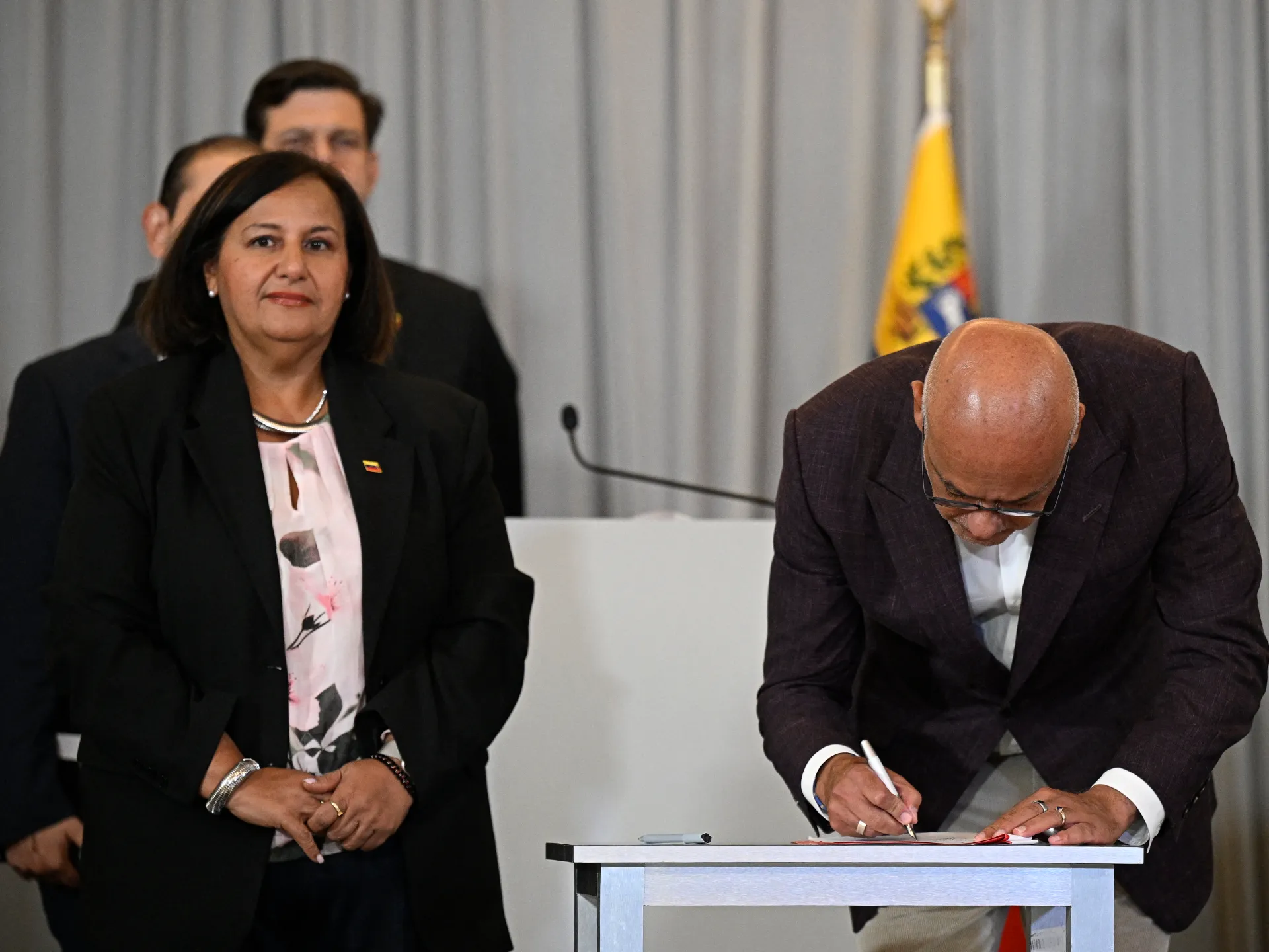 A woman stands beside a bald man signing a document at a table, with officials and a Venezuelan flag behind them.