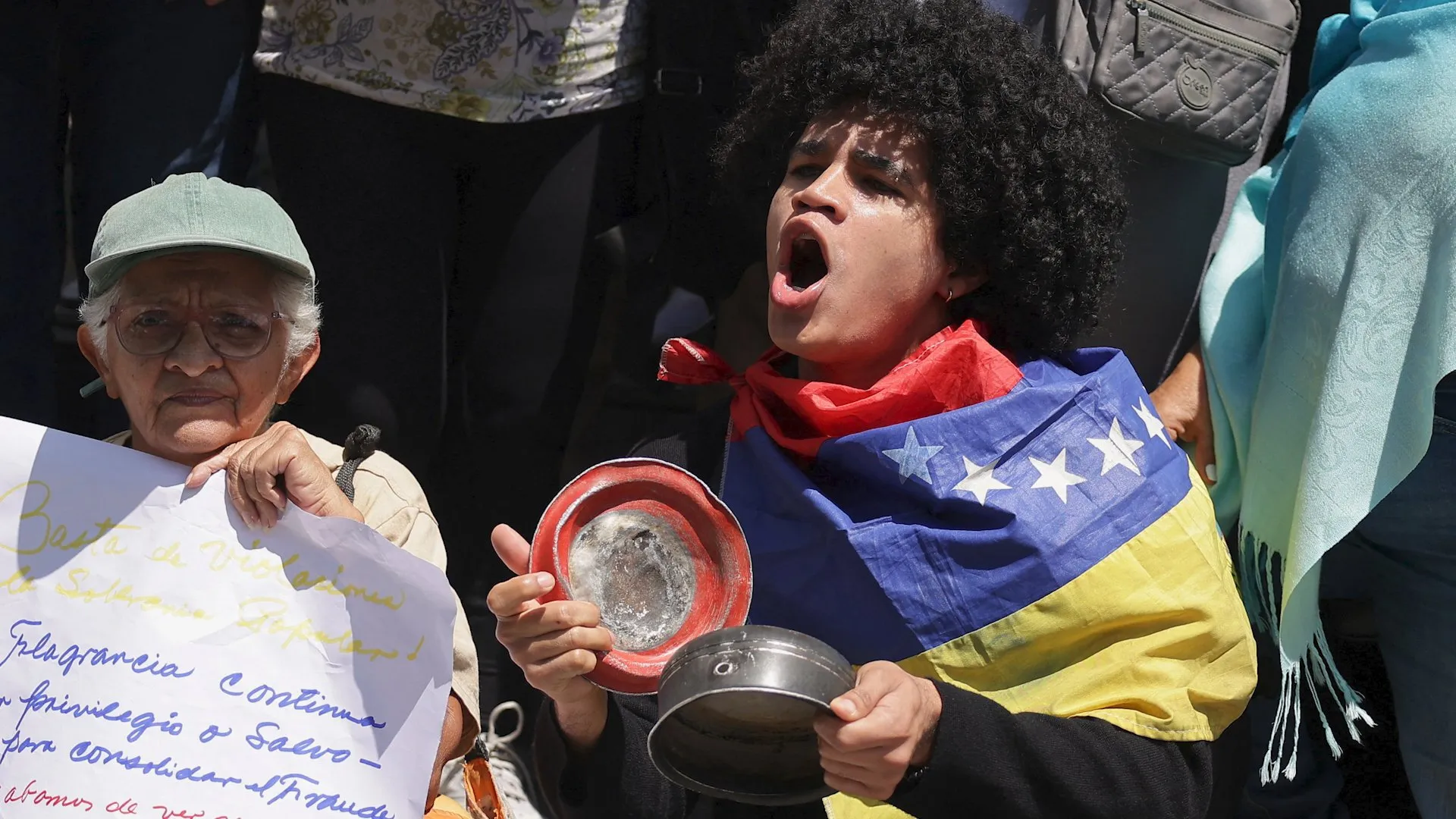 A person in a flag-themed outfit holds a small round object beside an older person holding a handwritten sign.