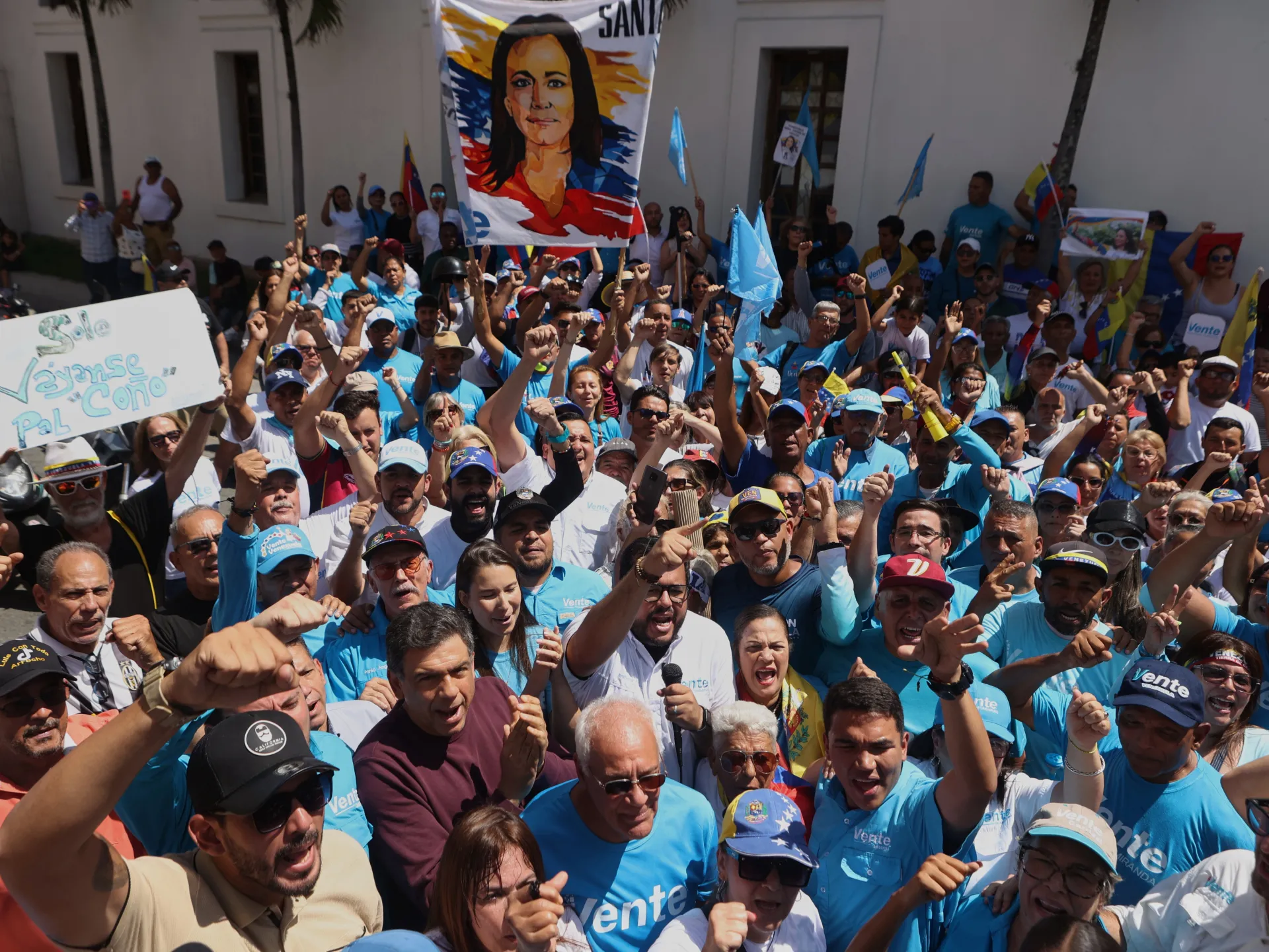 Crowd of people in blue shirts raise fists and flags beneath a large portrait banner.