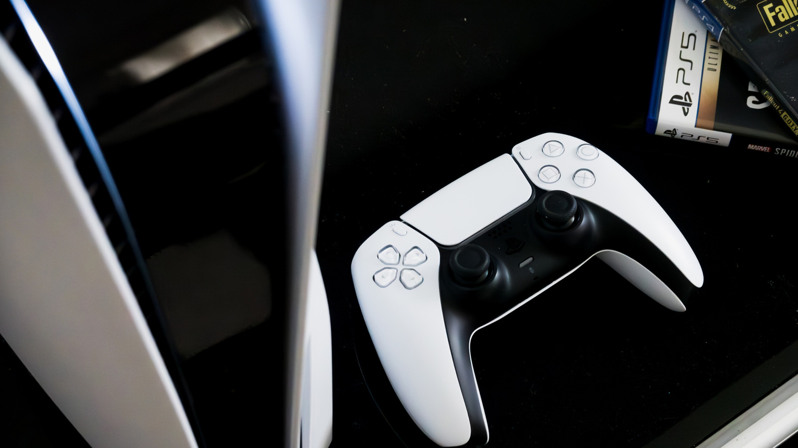 A white PlayStation 5 console stands beside a DualSense controller and partially visible game cases.