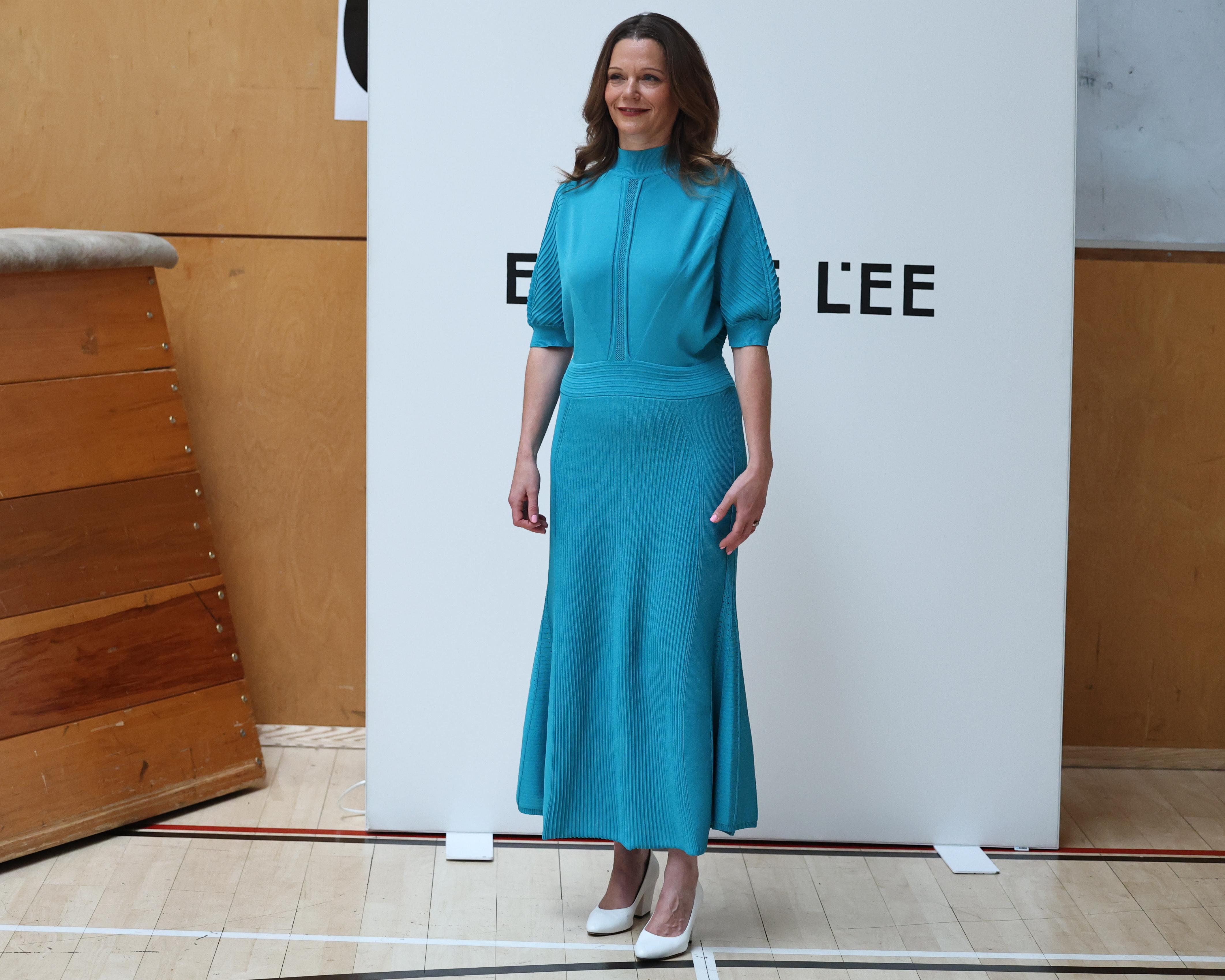 Woman in turquoise knit dress and white heels standing before a fashion show backdrop