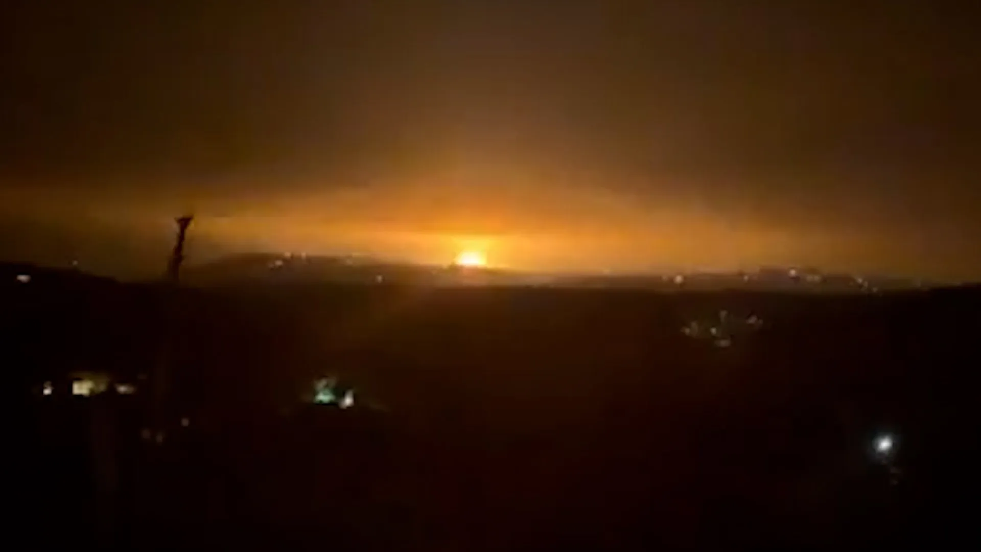 Bright orange flash illuminates a dark nighttime horizon beneath heavy clouds.