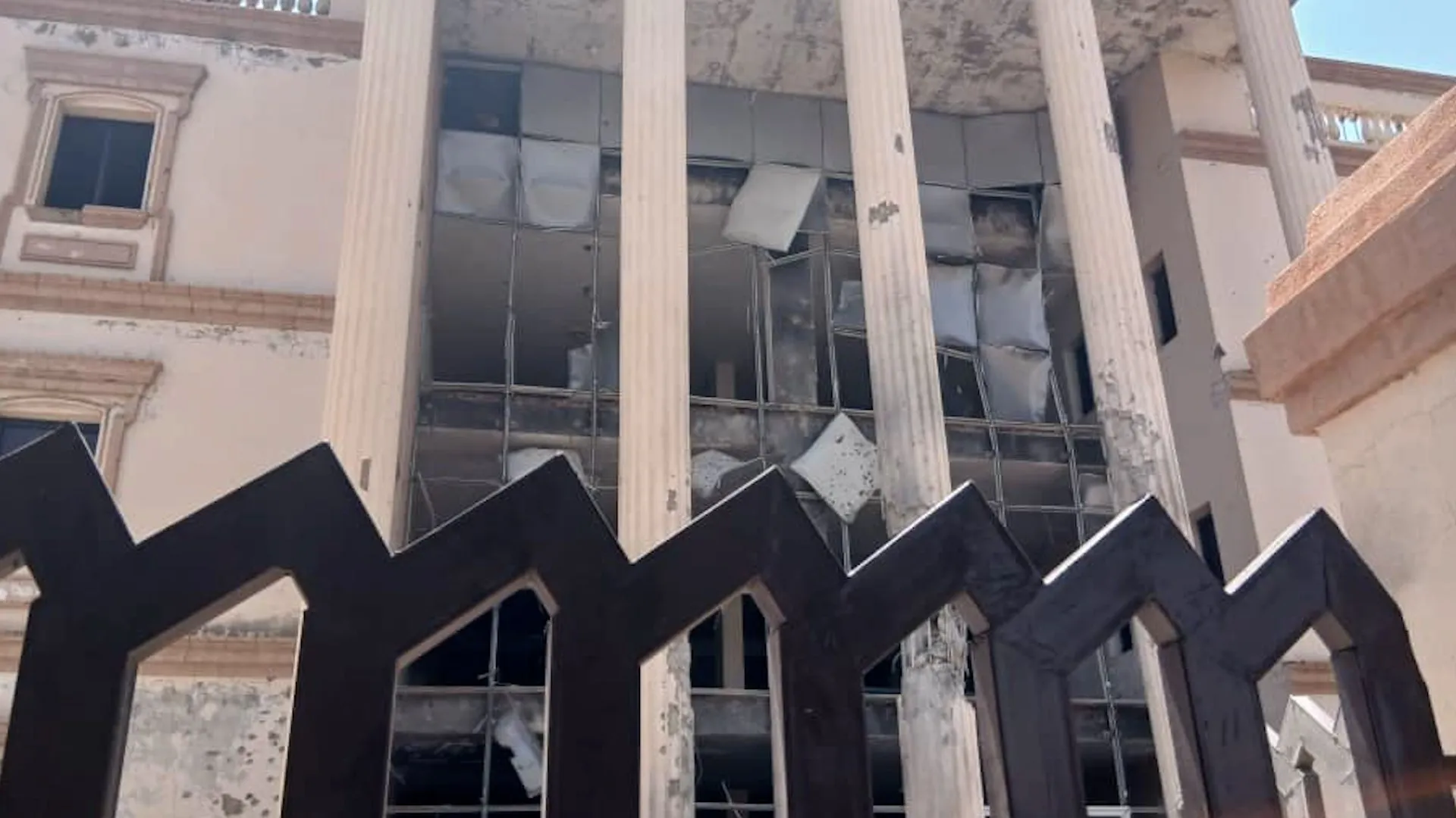 Damaged building with shattered windows, weathered columns, and debris visible across the façade.