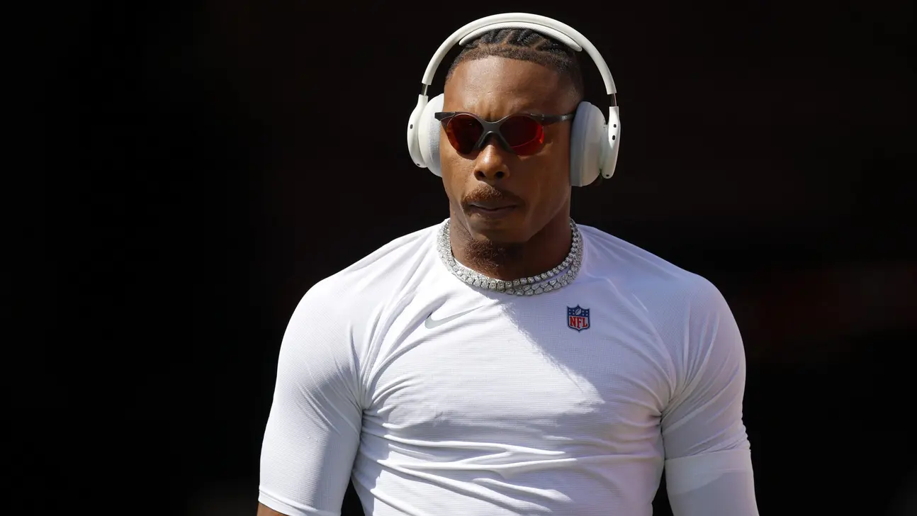 Man wearing white headphones, tinted sunglasses, a white NFL shirt, and a chain against a dark background.