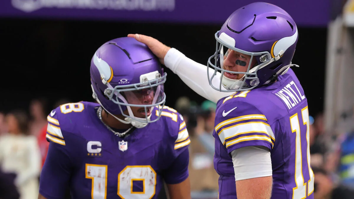 Two Minnesota Vikings players in purple uniforms stand together, one patting the other's helmet.