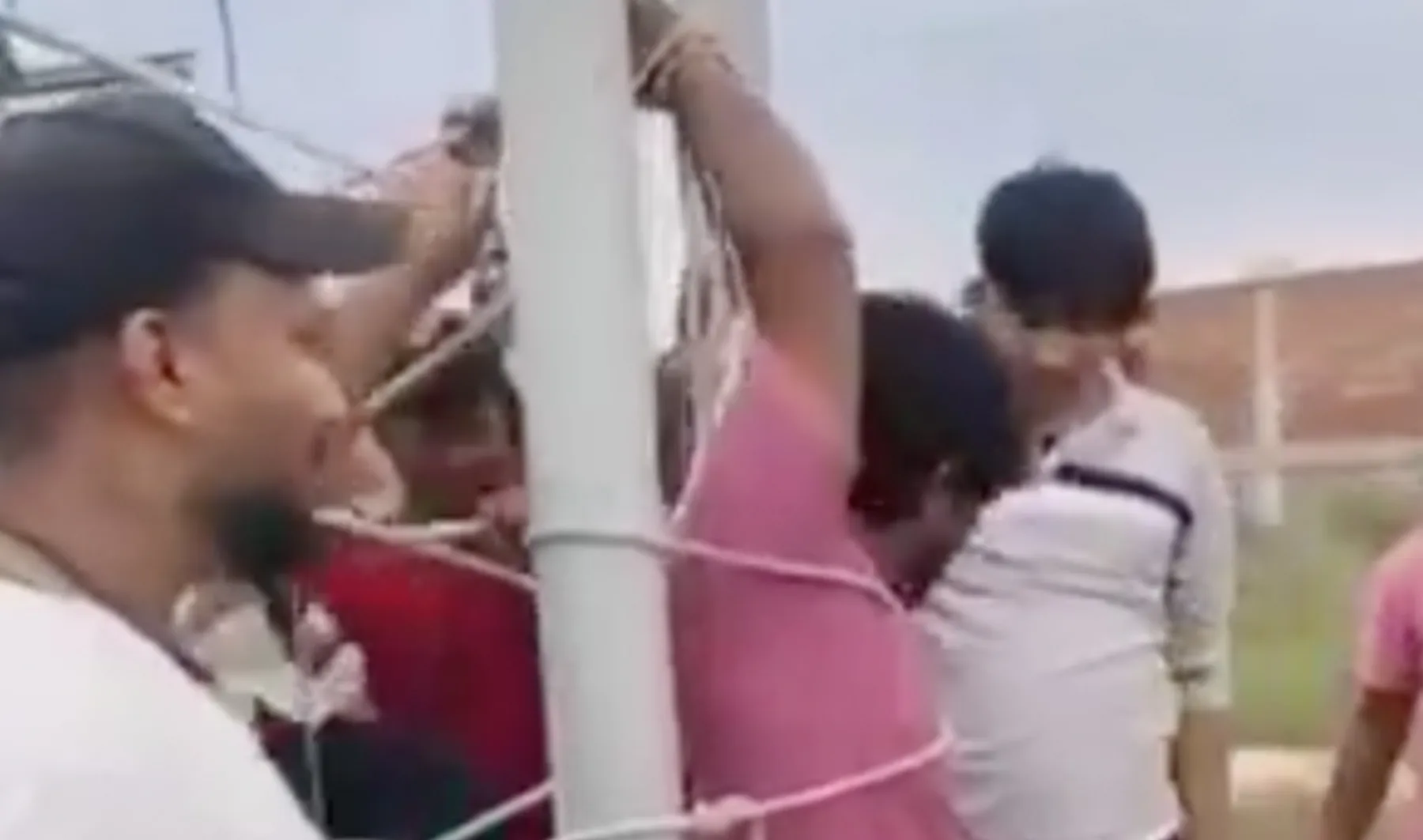Several people restrain a man in a pink shirt against a white cell tower structure.