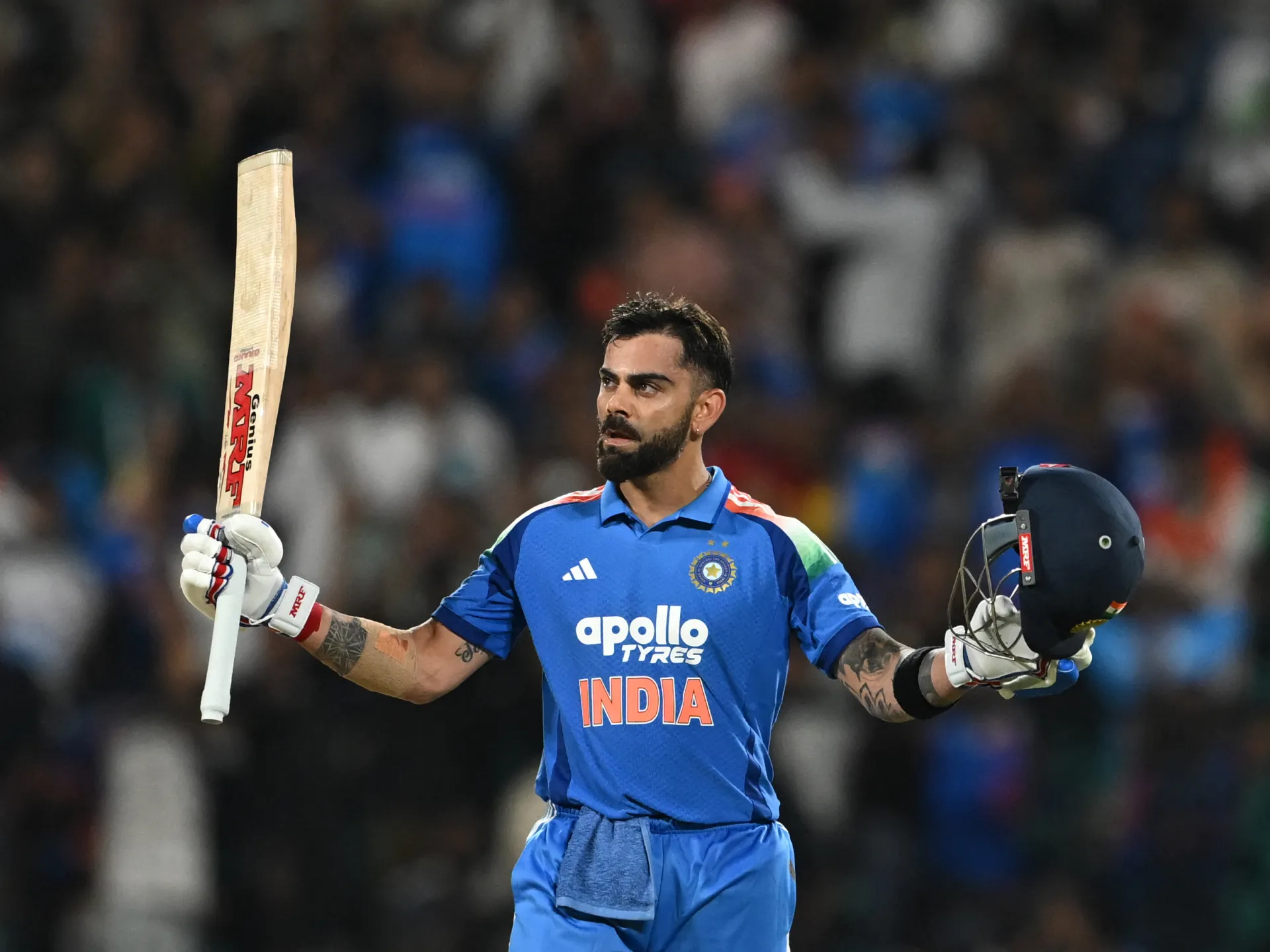 Indian cricketer in blue raises his bat and holds his helmet during a match.