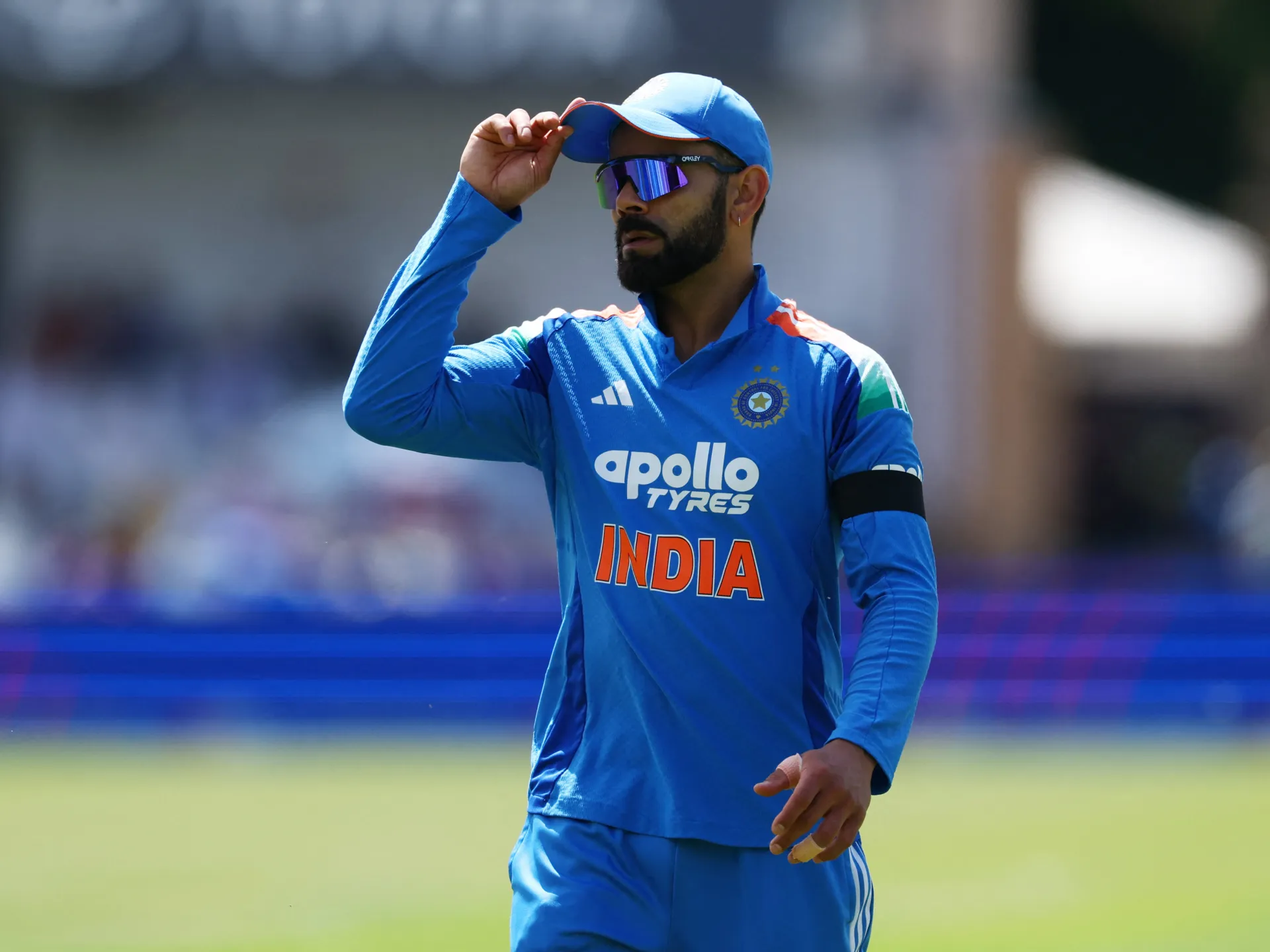 Cricketer in a blue India uniform adjusts his cap while wearing mirrored sunglasses on the field.