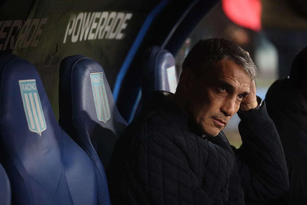 Man seated on a football team bench, resting his head on one hand beside empty blue seats.