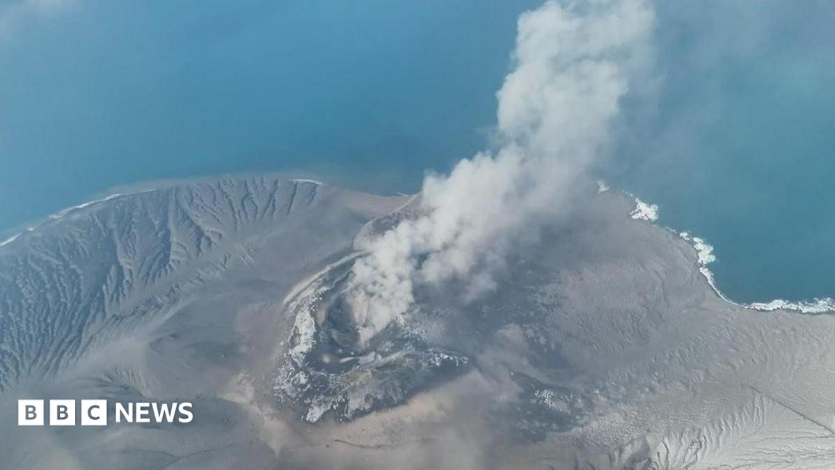 Aerial view of a smoking volcanic crater on a barren island surrounded by blue ocean