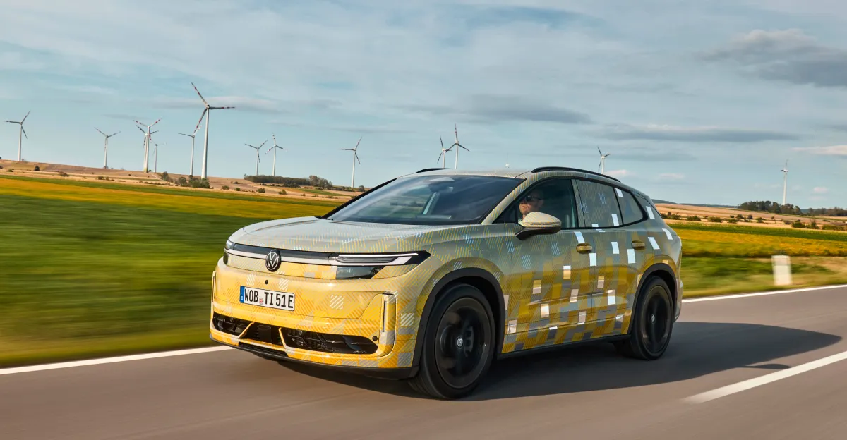 Camouflaged Volkswagen electric SUV driving past wind turbines on a rural road