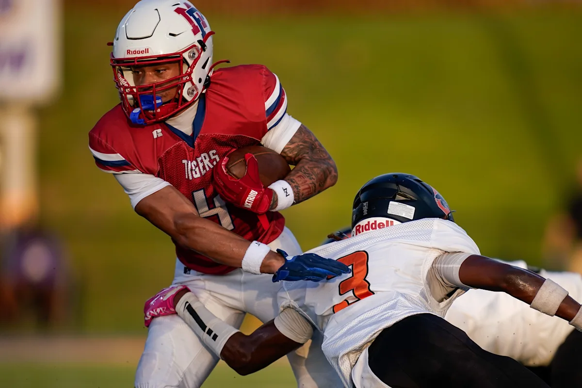 Football player in red jersey carries ball while evading a defender in white uniform