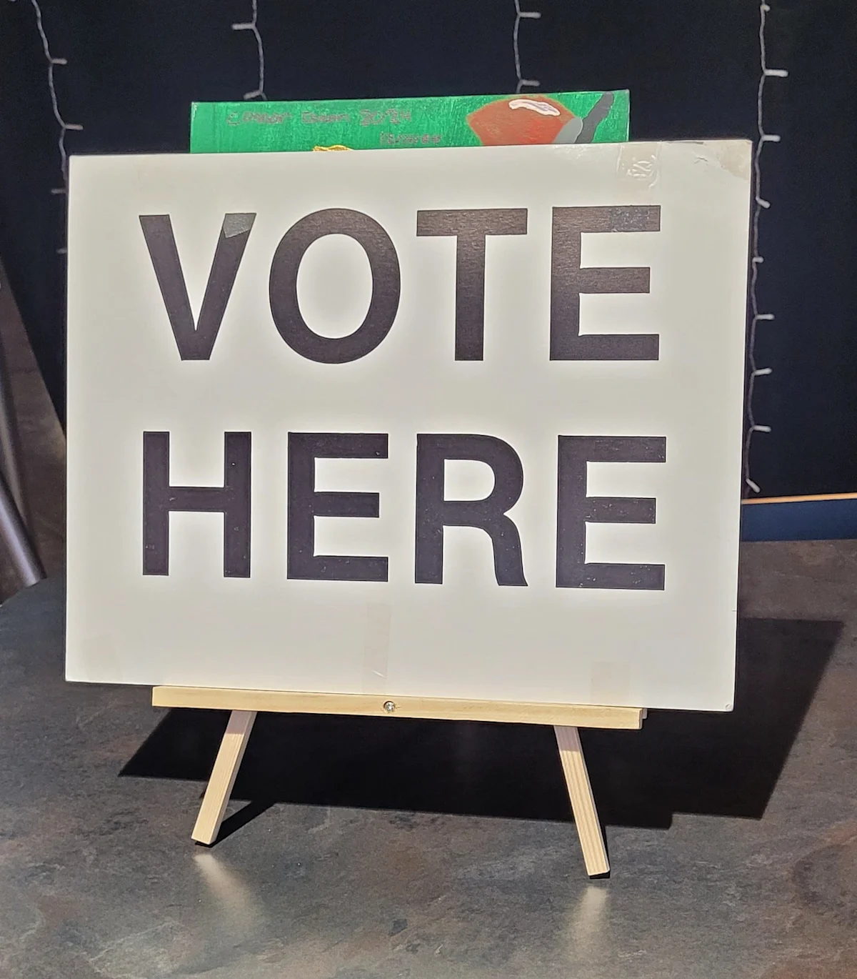 White easel sign with large black letters reading “VOTE HERE” against a dark background.