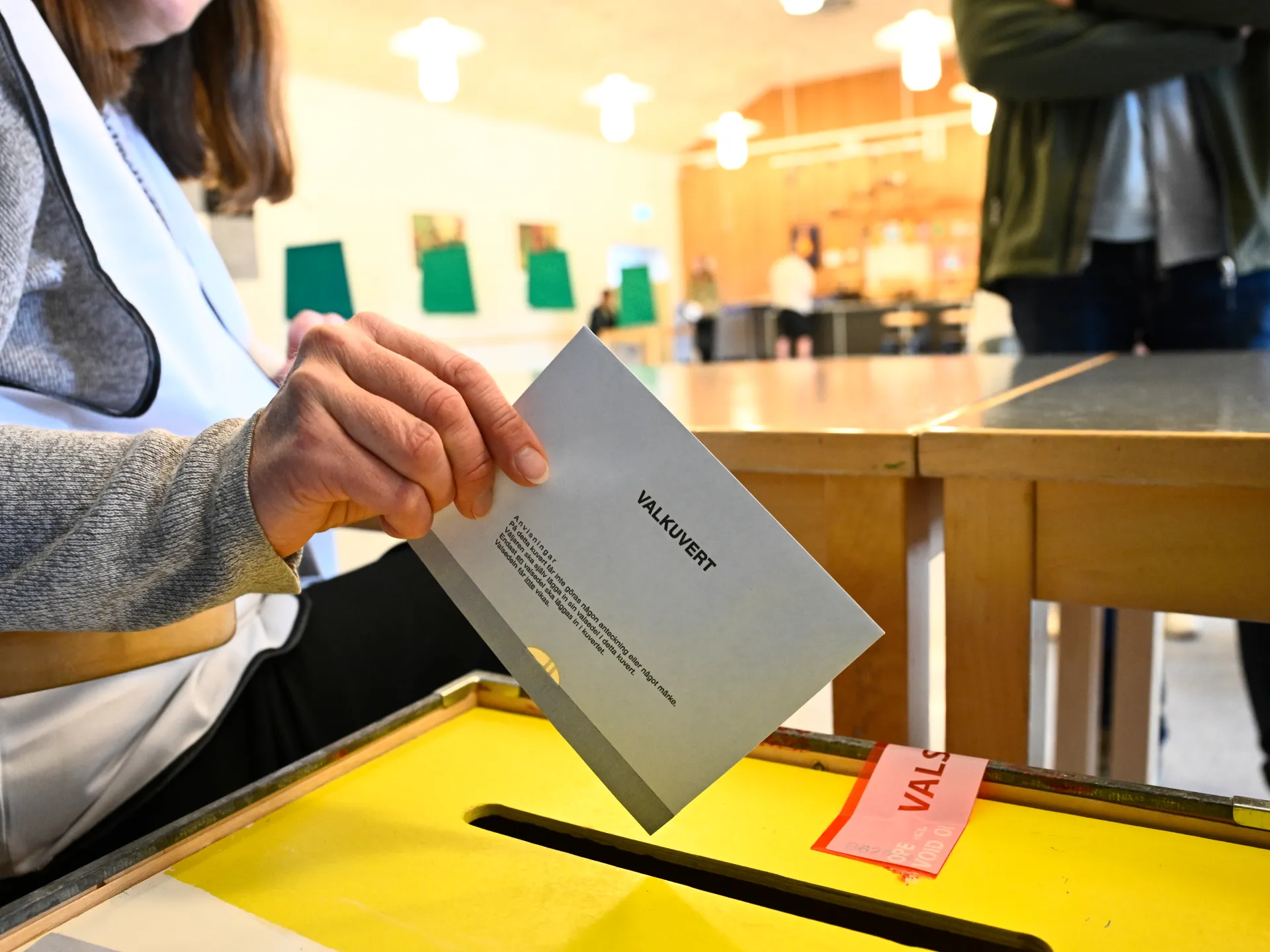 Voter inserts a ballot envelope into a yellow ballot box at a polling station.