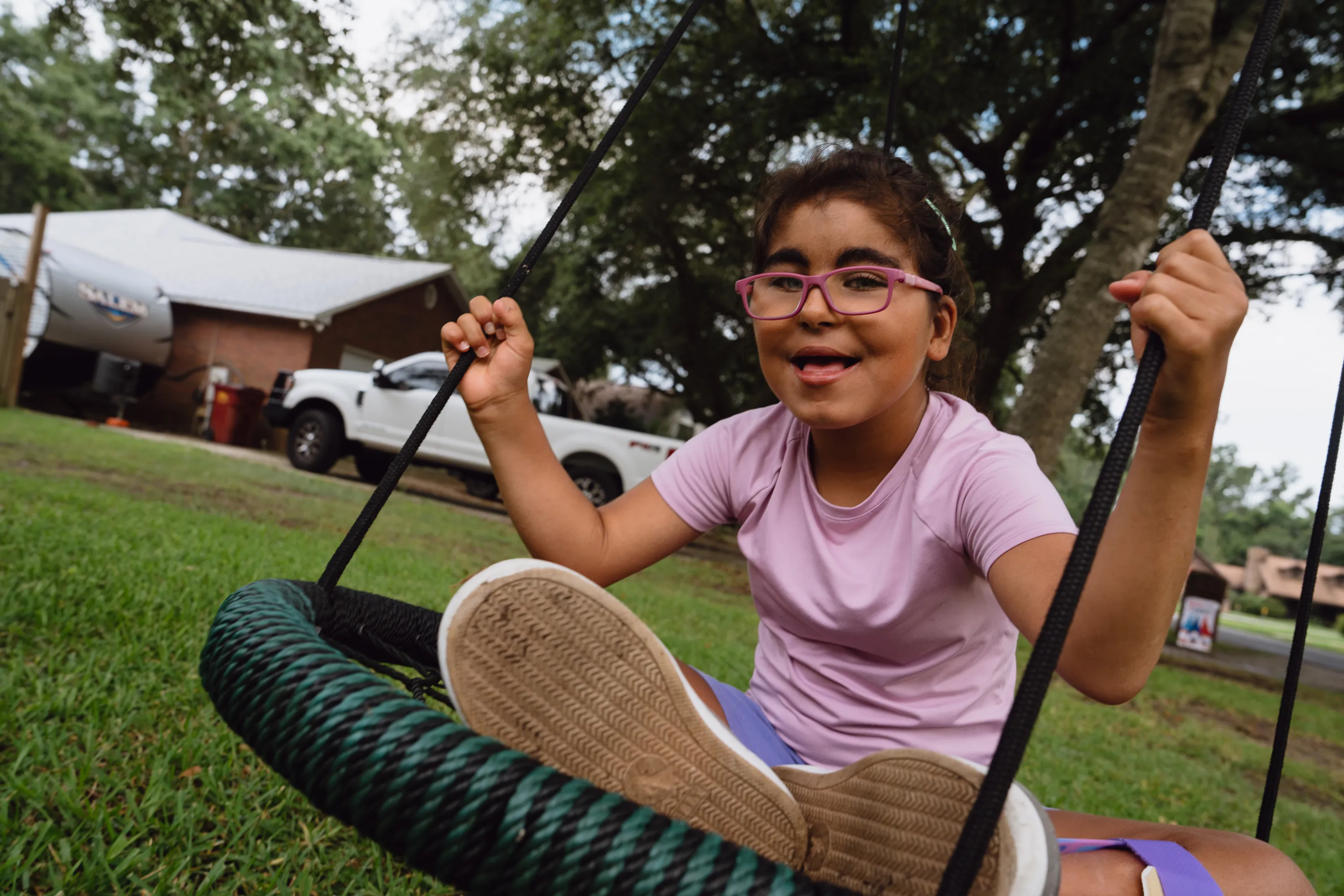 Child wearing pink glasses swings outdoors, holding swing ropes with a sneaker raised toward camera.