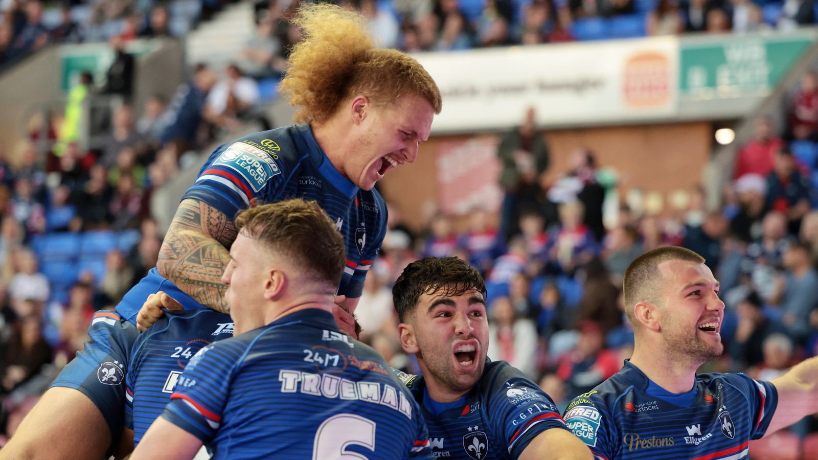 Blue-shirted rugby players celebrate together on the pitch in front of a crowded stand.