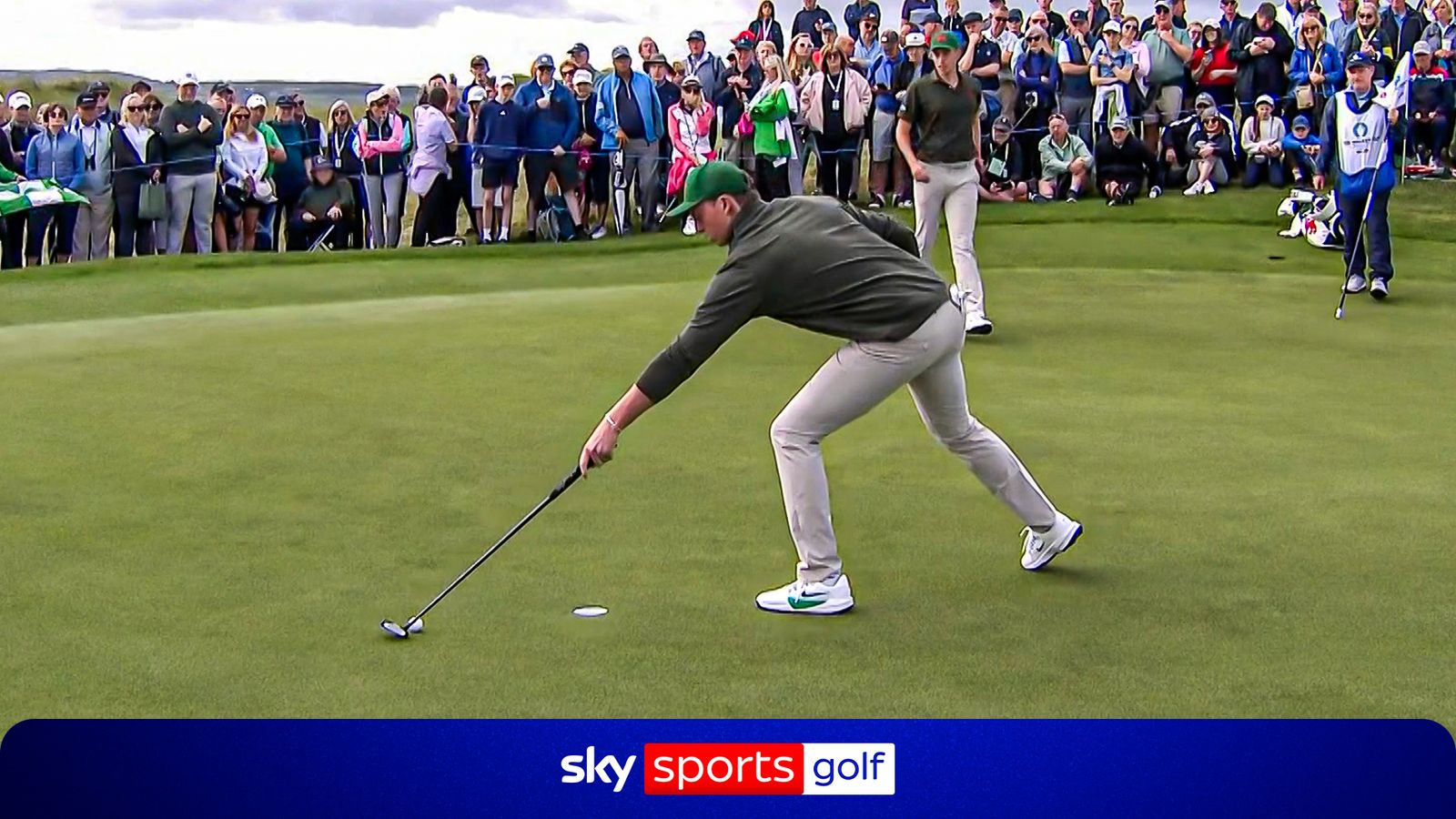 Golfer bends to retrieve a ball near the hole as spectators watch on a putting green.