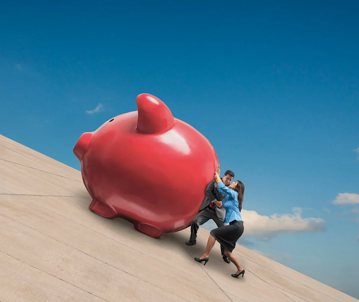 Large red piggy bank and two businesspeople struggle up a steep concrete slope beneath blue sky