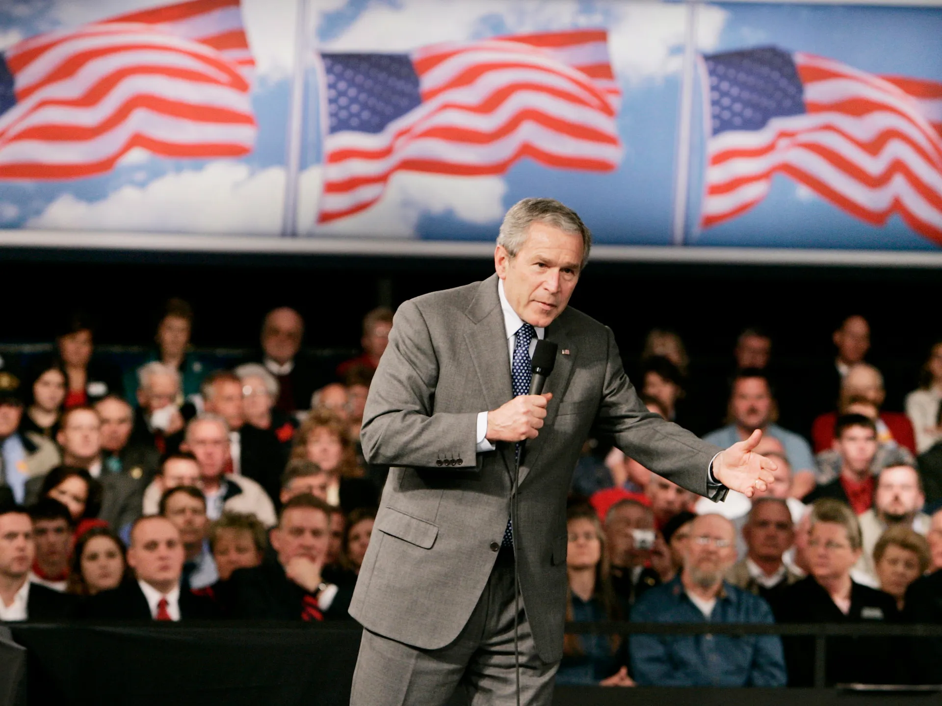 Suited speaker addresses a seated audience before several American flags.