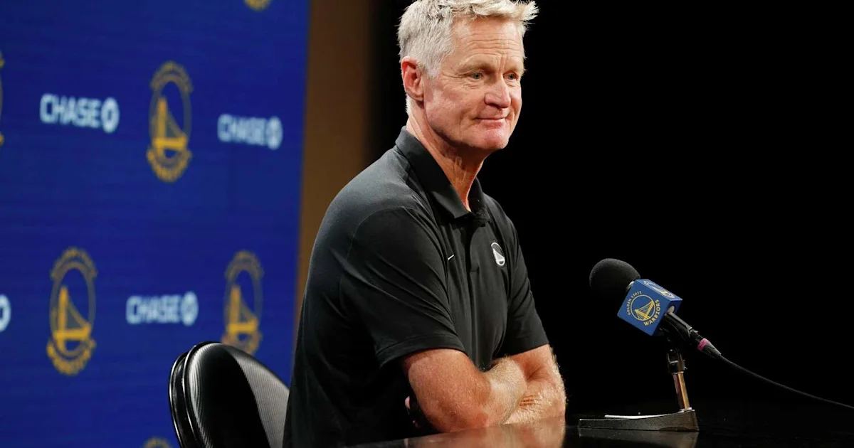 A gray-haired man in a black polo sits at a press conference beside a microphone.