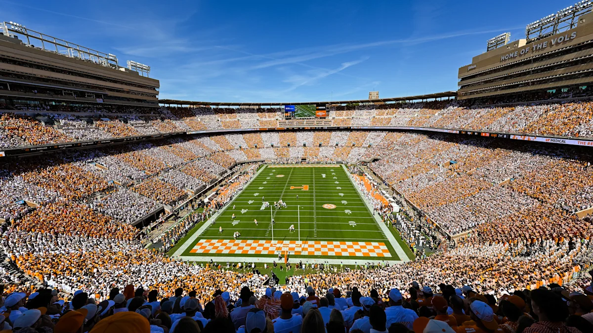 A packed football stadium surrounds the field beneath a clear blue sky during a game.
