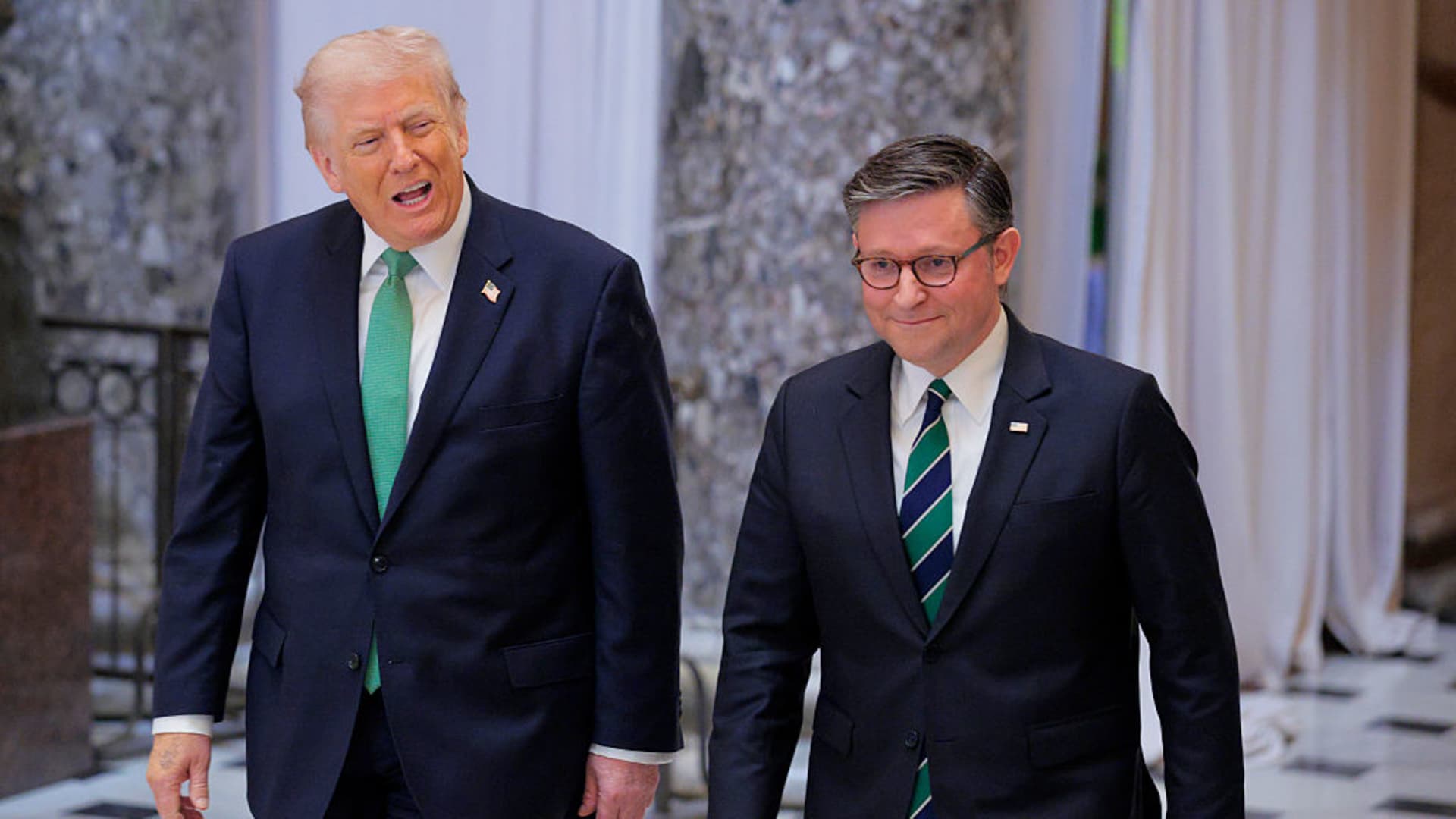 Two men in dark suits walk indoors, with one speaking and the other smiling.