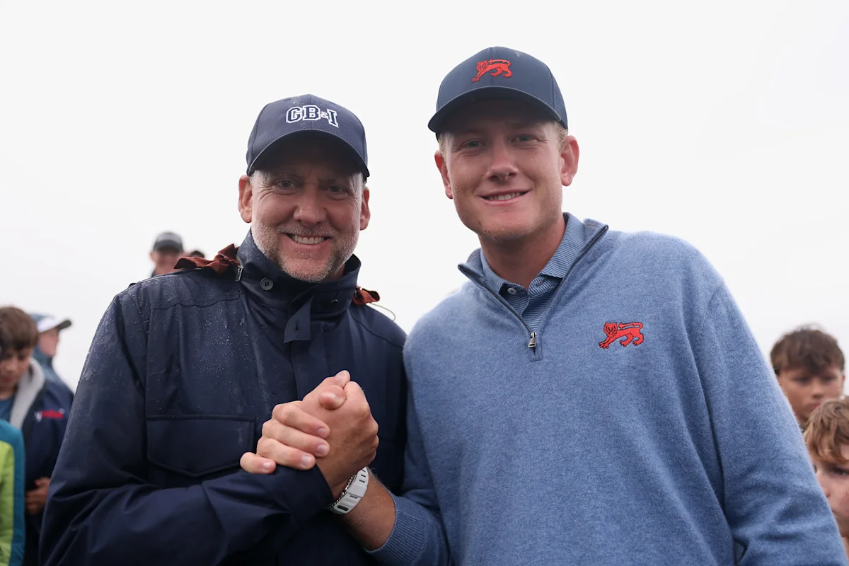 Two men in caps and sportswear smile and shake hands outdoors as spectators gather behind them.