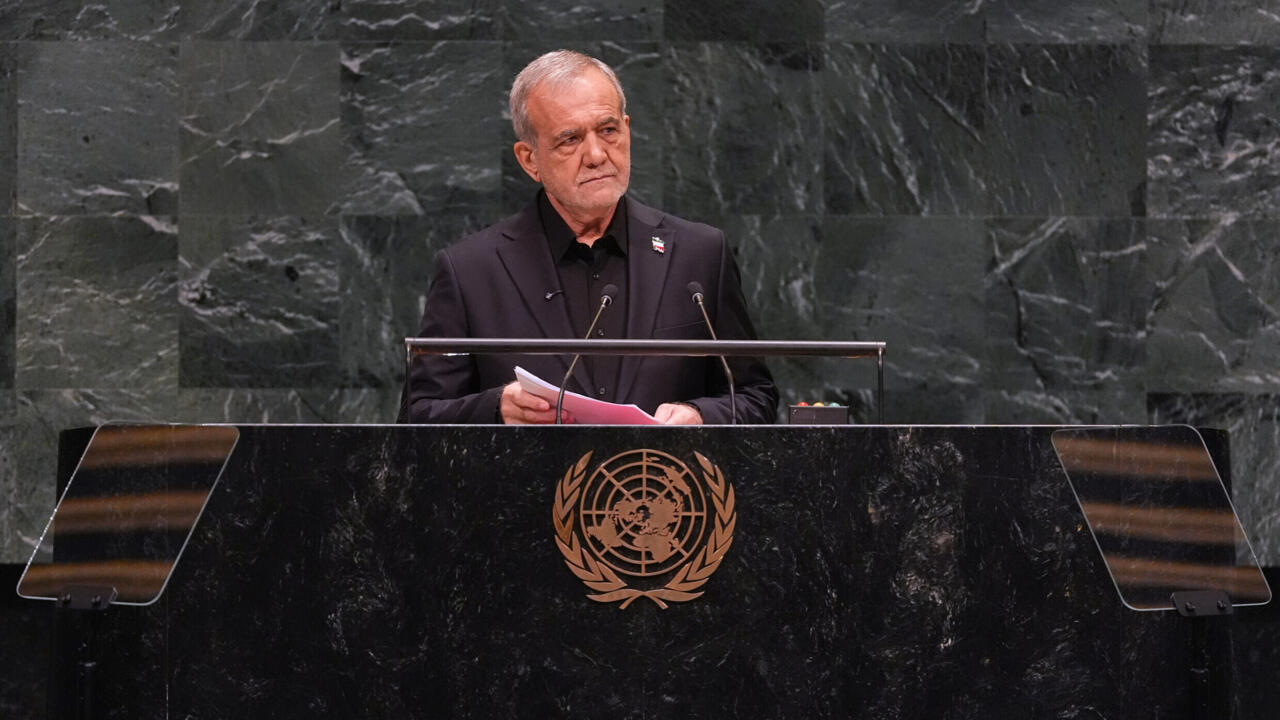 A gray-haired man in a dark suit speaks from behind a lectern bearing the United Nations emblem.