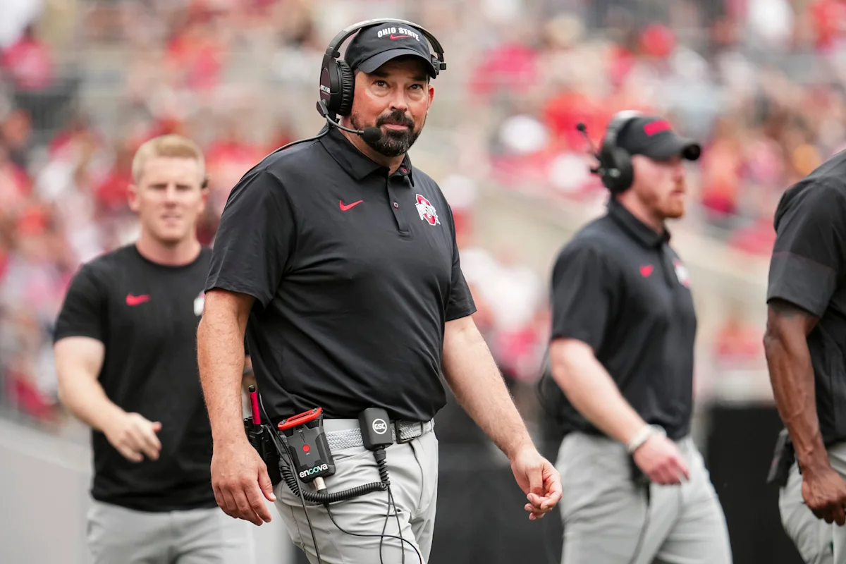 Football coach wearing a headset and Ohio State apparel walks along the sideline.