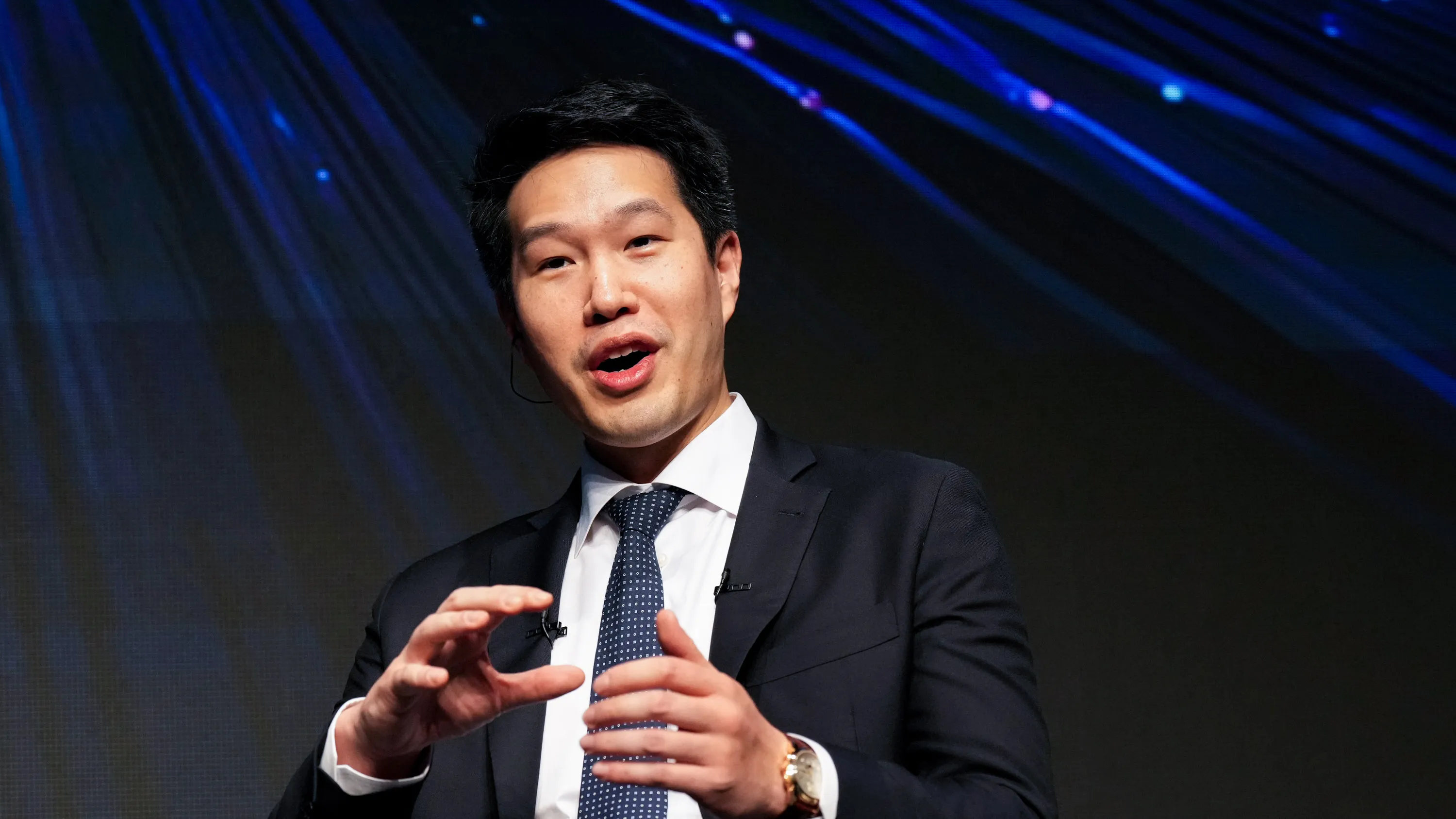 A suited speaker gestures while addressing an audience beneath blue stage lights.