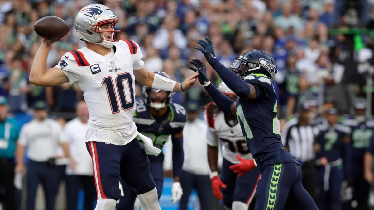 A Patriots player throws a football as a Seahawks player raises an arm nearby on the field.