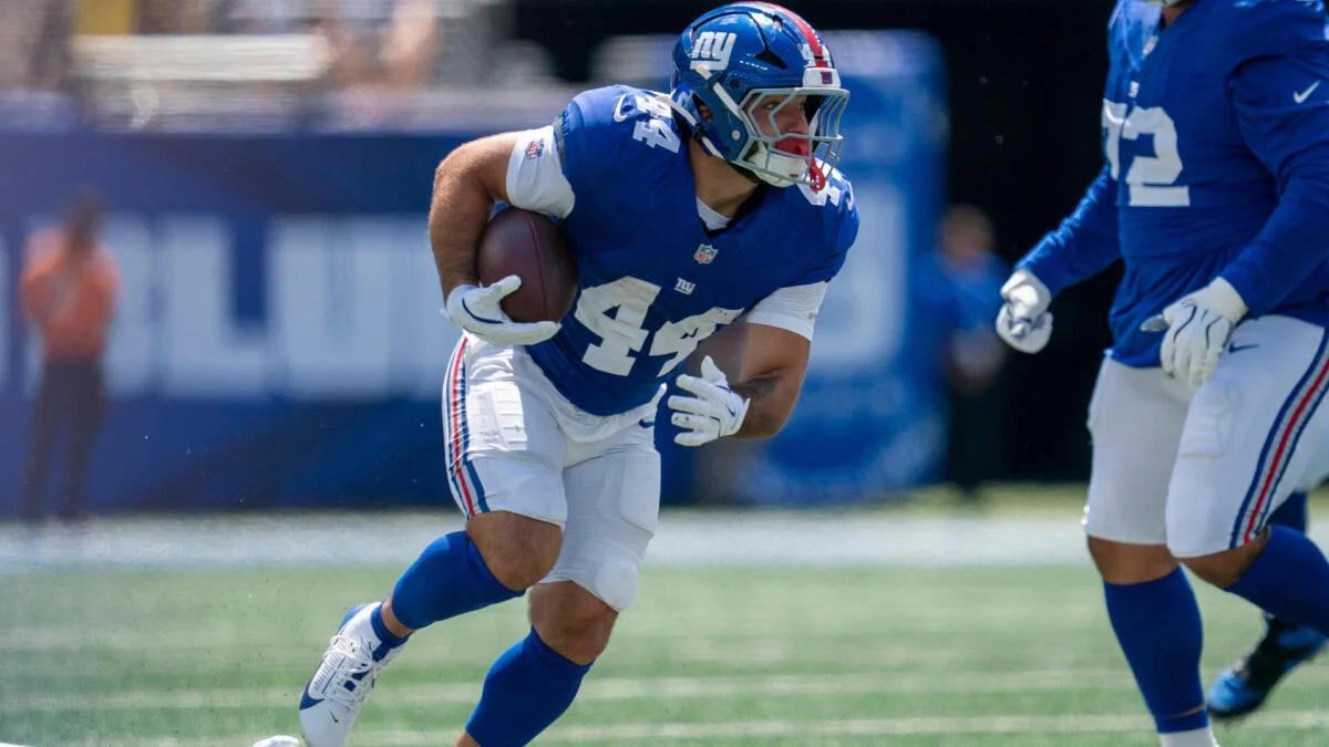 A New York Giants player in a blue jersey runs with the football during practice.