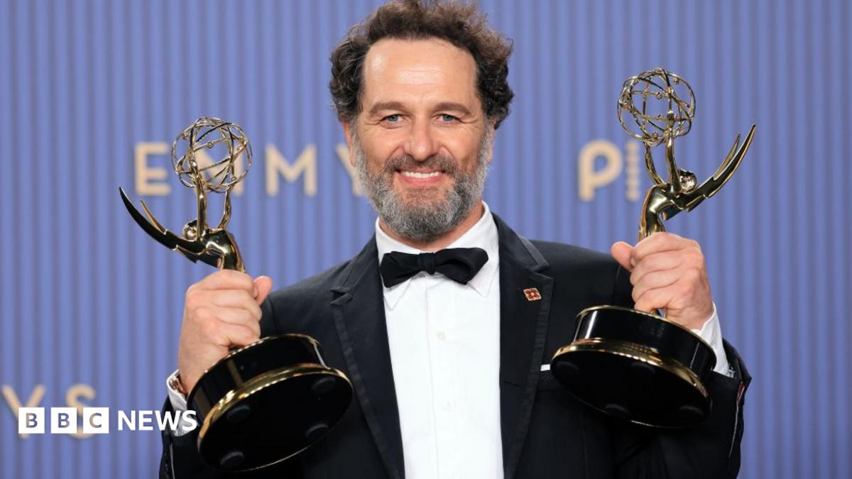 Smiling bearded man in tuxedo holds two Emmy statuettes at an awards ceremony.