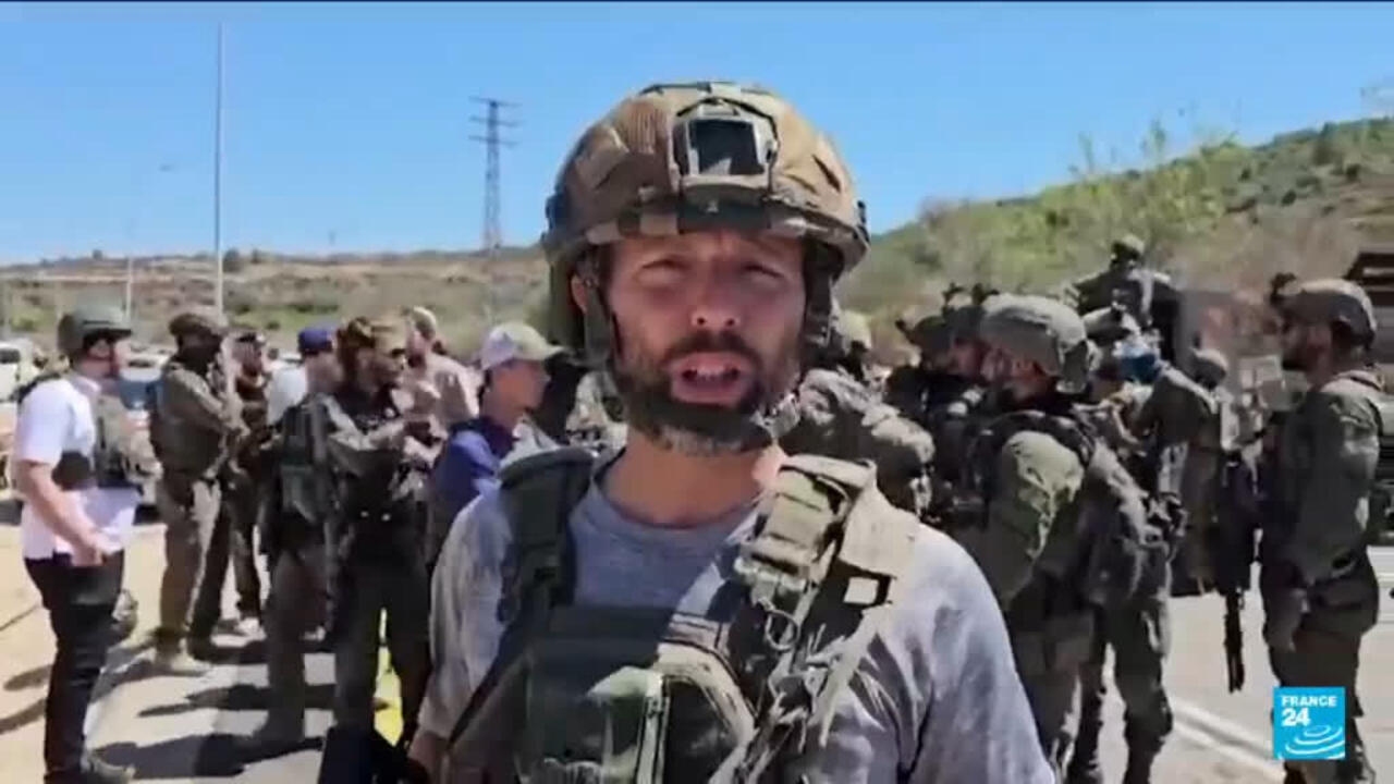 Bearded man in military helmet and tactical vest stands before armed personnel outdoors.