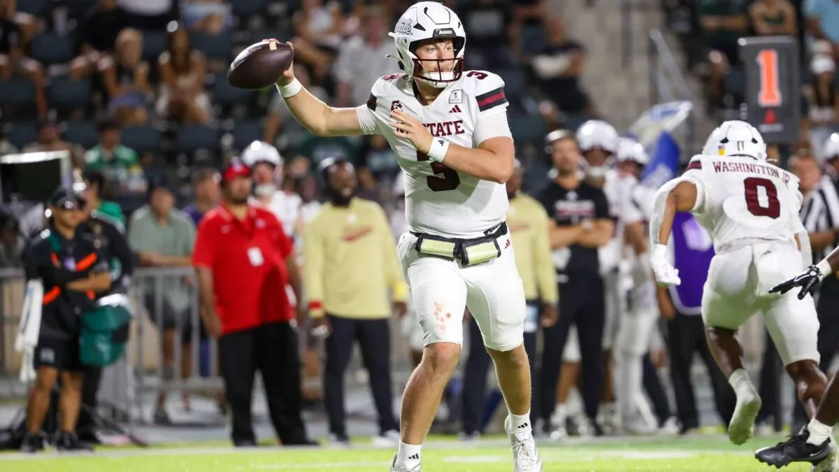 A helmeted quarterback in a white uniform prepares to throw as players and spectators fill the background.