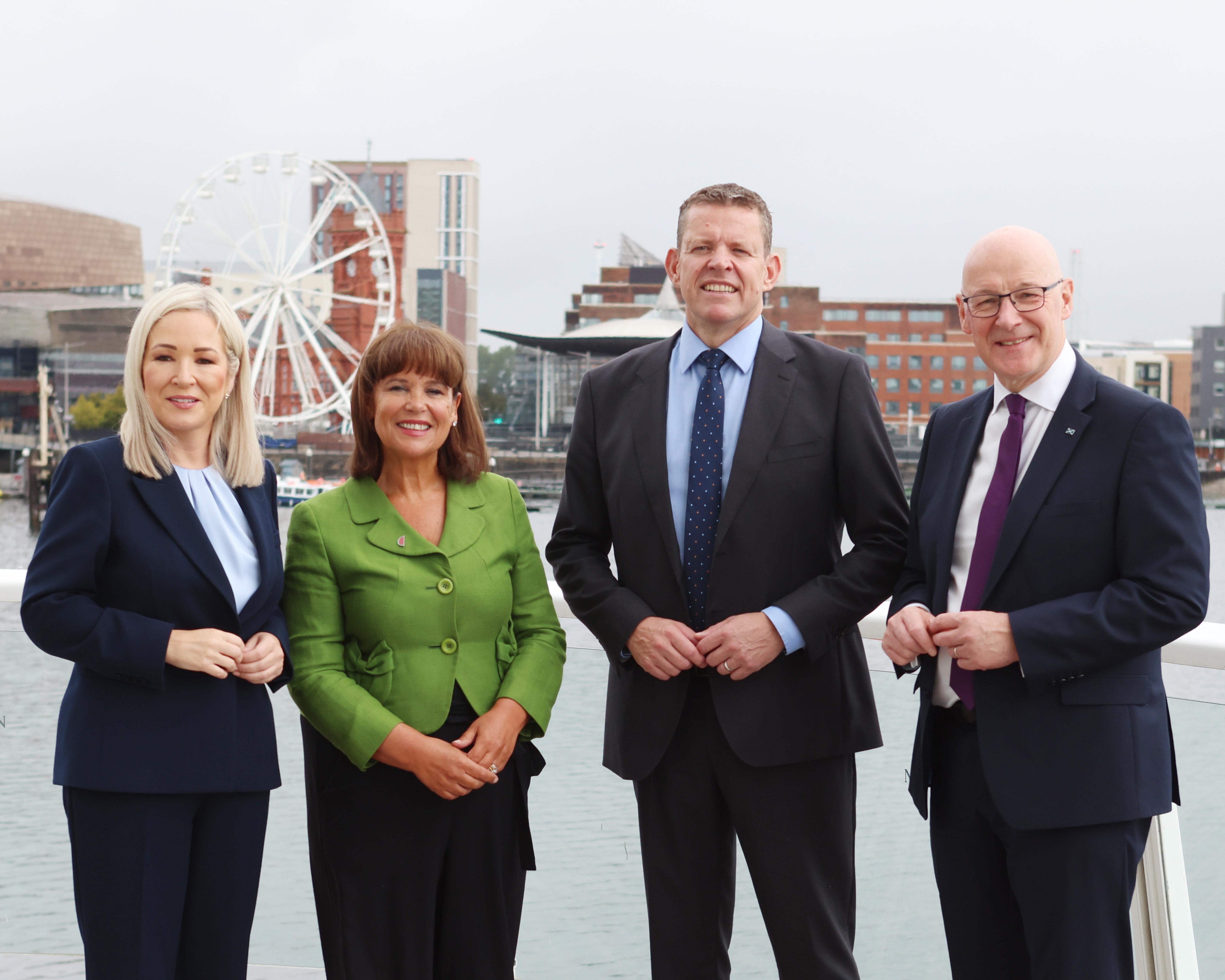 Four formally dressed officials pose beside waterfront with a Ferris wheel and city buildings behind them.