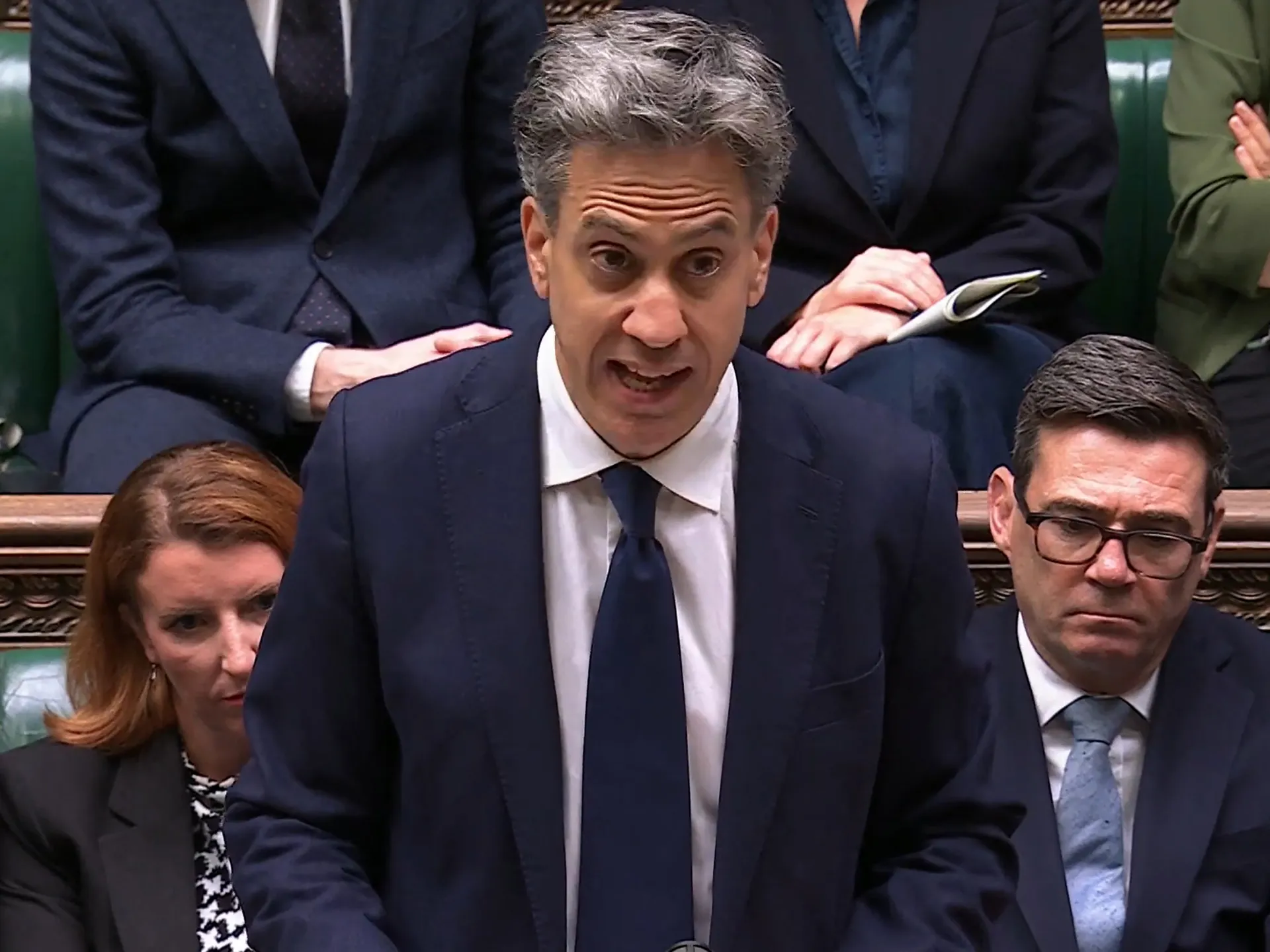 Grey-haired man in a dark suit speaks in Parliament, with seated colleagues behind him.