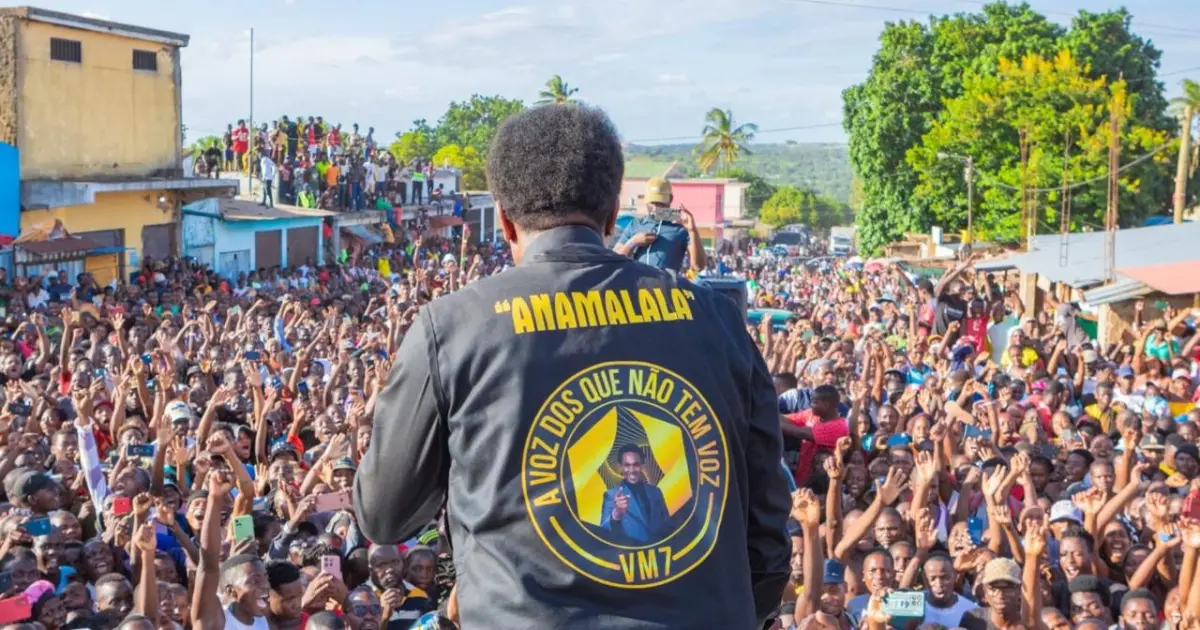 A speaker in a black jacket addresses a large cheering crowd from an elevated platform.