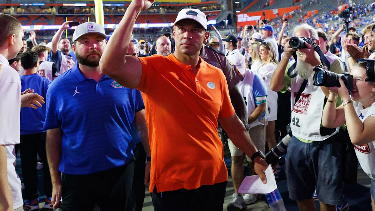 Man in orange polo and white cap walks through a crowd as photographers take pictures.