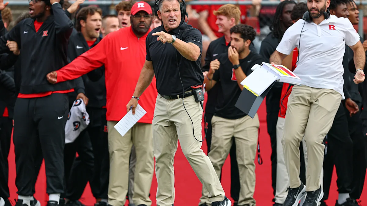 Several football coaches stand on the sideline, with one pointing and others holding papers.