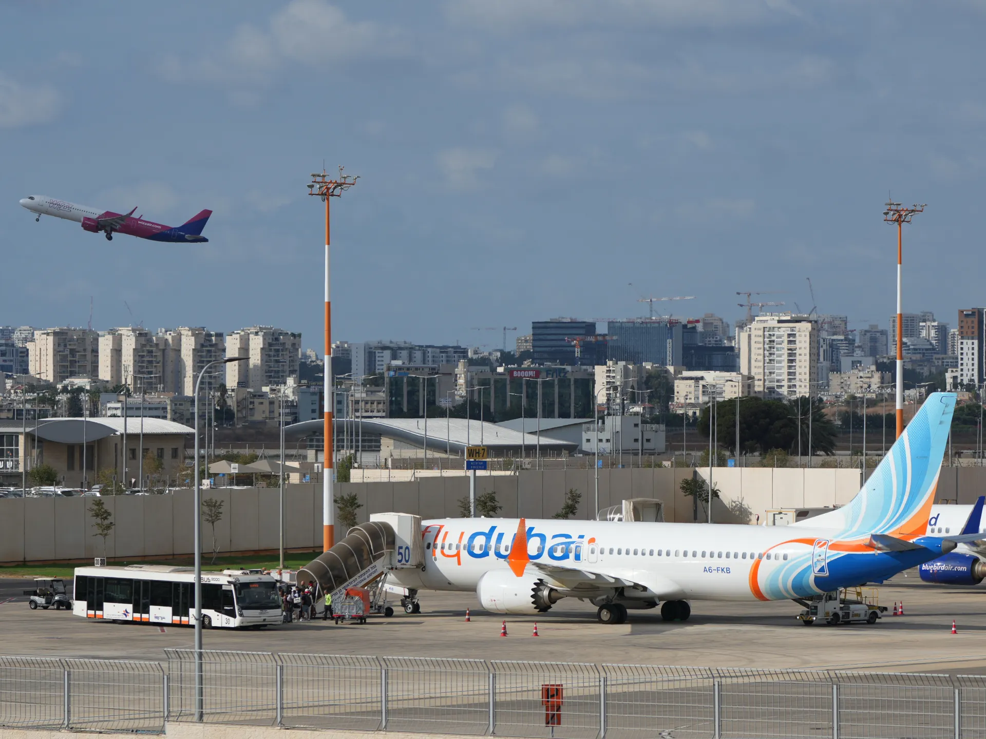 Flydubai passenger plane parked at an airport as another aircraft climbs overhead.