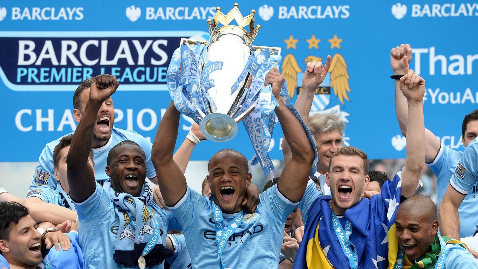 Manchester City players celebrate with the Premier League trophy amid blue banners and raised arms.