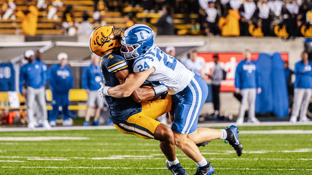 A football player in blue and white tackles a player in navy and gold on the field.