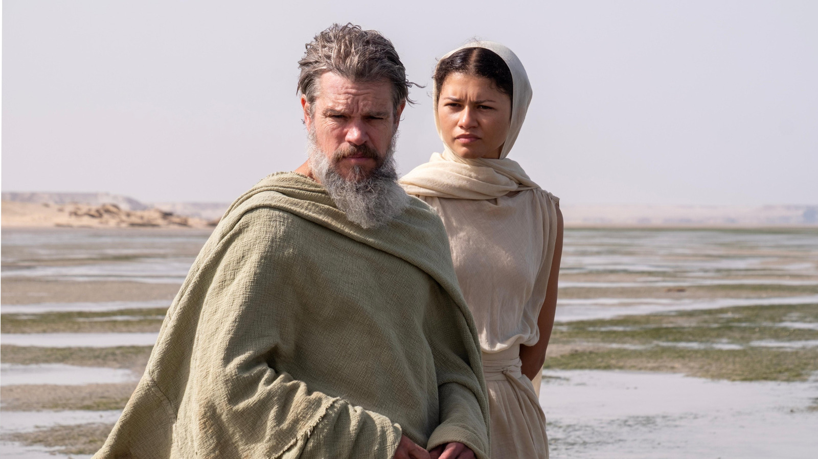 Bearded man and headscarf-wearing woman stand together beside a shallow, rippled shoreline.