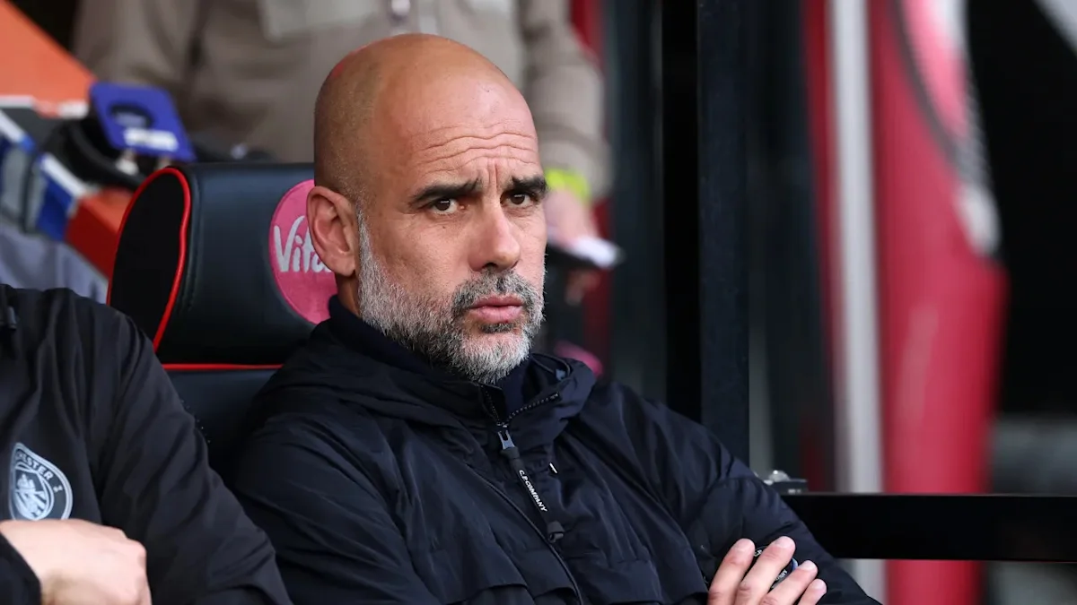Bald, bearded man in a black jacket sits with arms crossed on a stadium bench.