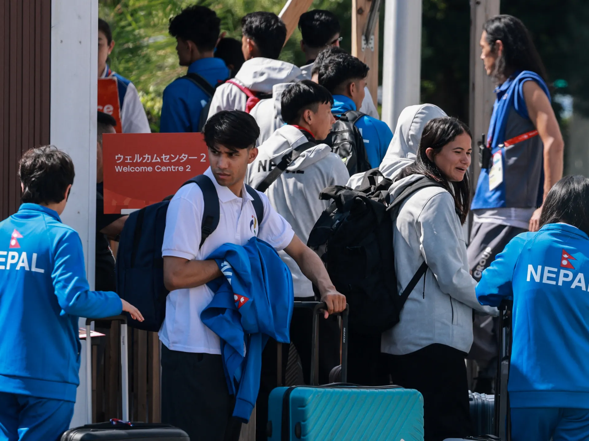 Athletes and travelers with luggage gather near a Welcome Centre 2 entrance.
