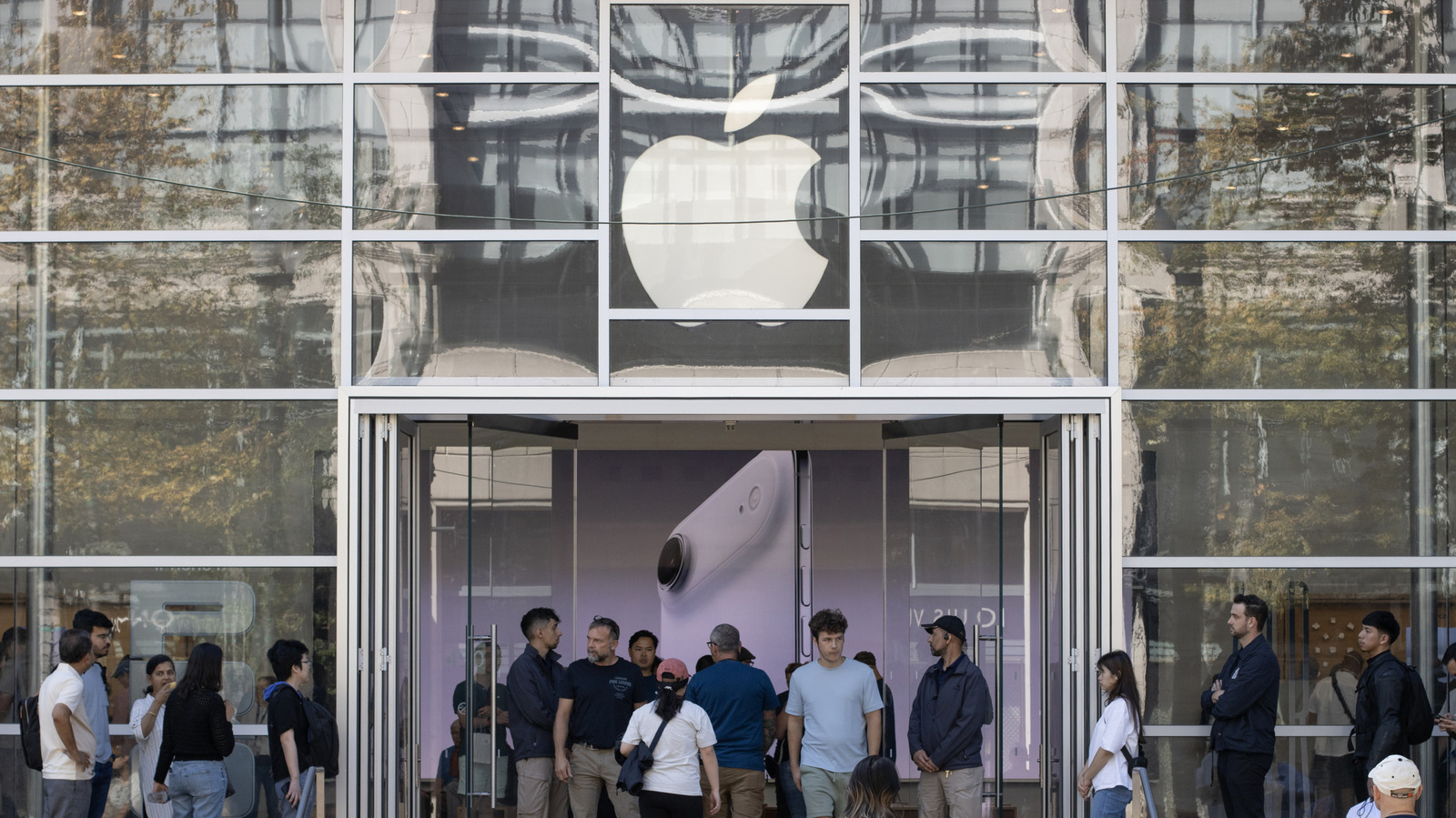 People stand outside an Apple Store entrance beneath a large illuminated Apple logo.