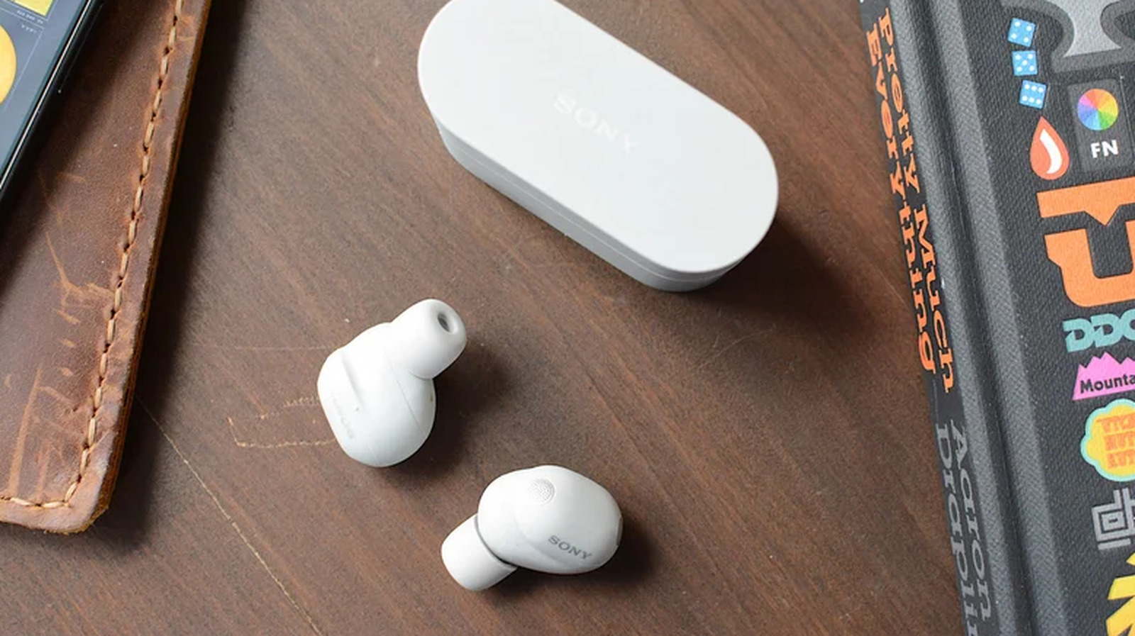 Two white Sony wireless earbuds and charging case on a wooden desk beside a book