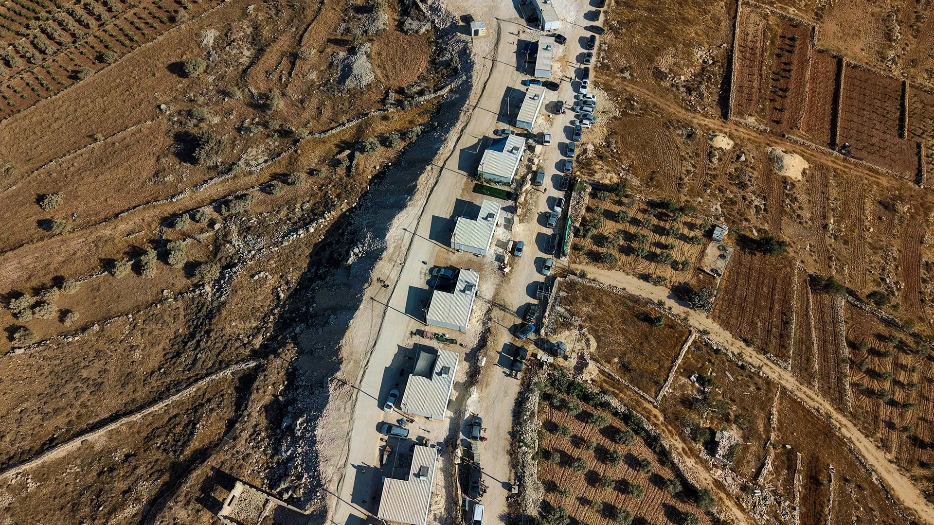 Aerial view of farm plots, a rocky streambed, dirt road, buildings, and parked vehicles.