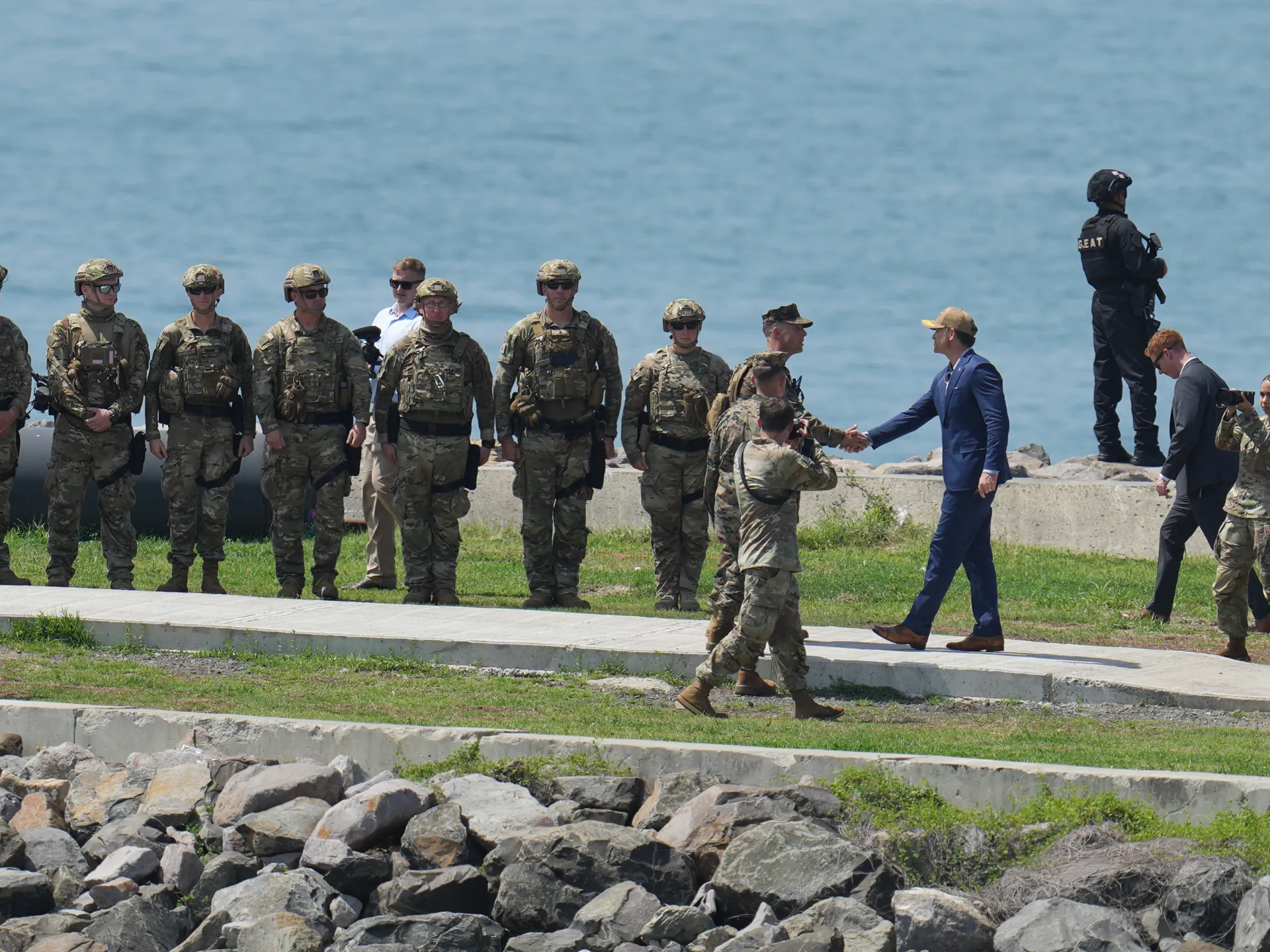 Military personnel stand near a waterfront as a suited official shakes hands with a soldier.